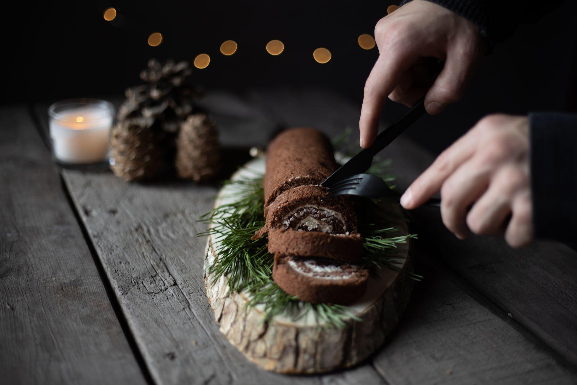 A person is slicing a chocolate yule log cake decorated with pine sprigs on a rustic wooden board, with blurred fairy lights, a candle, and pinecones in the background.