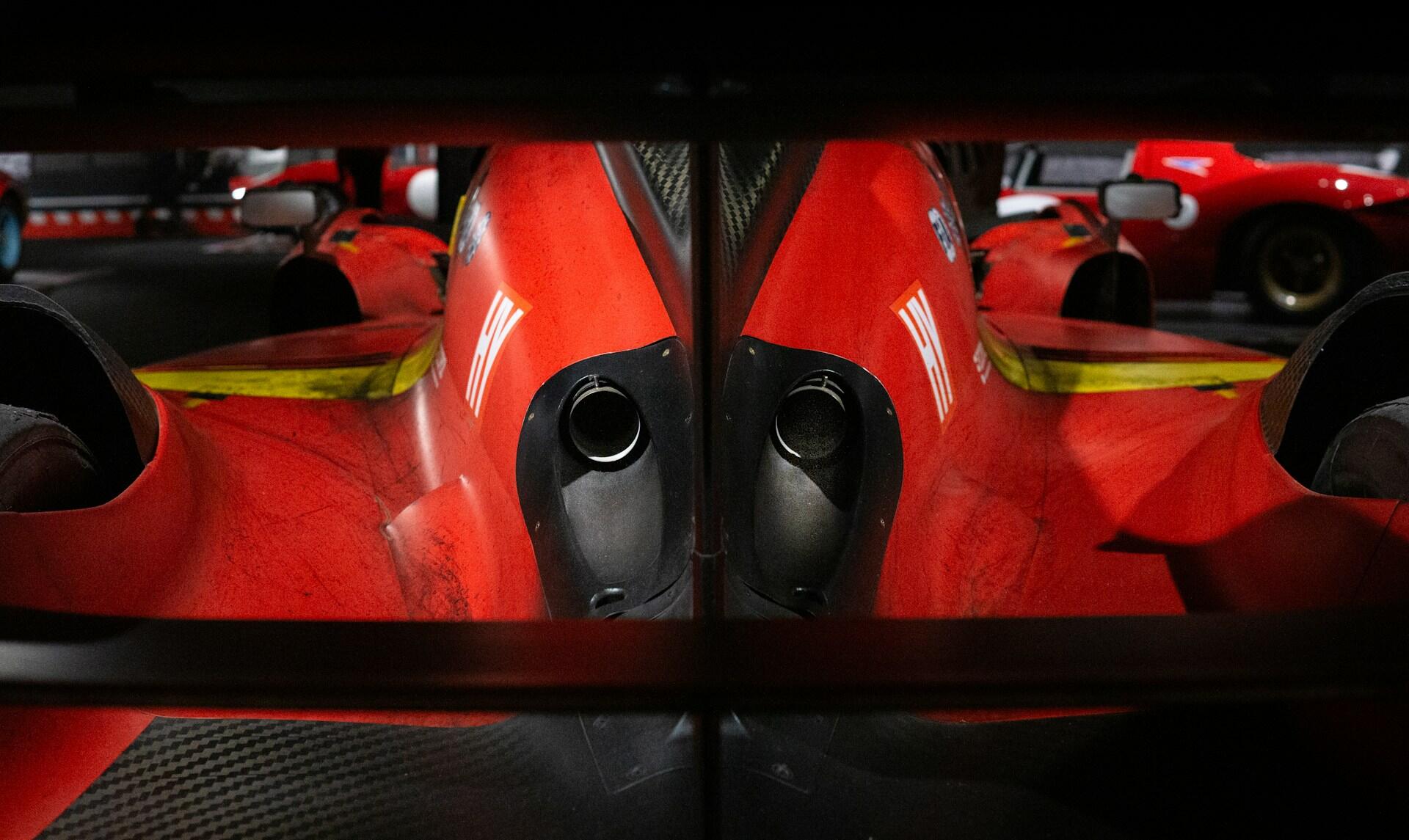 Rear close-up of a red racing car showing central exhaust pipes and aerodynamic design details in a dimly lit garage.