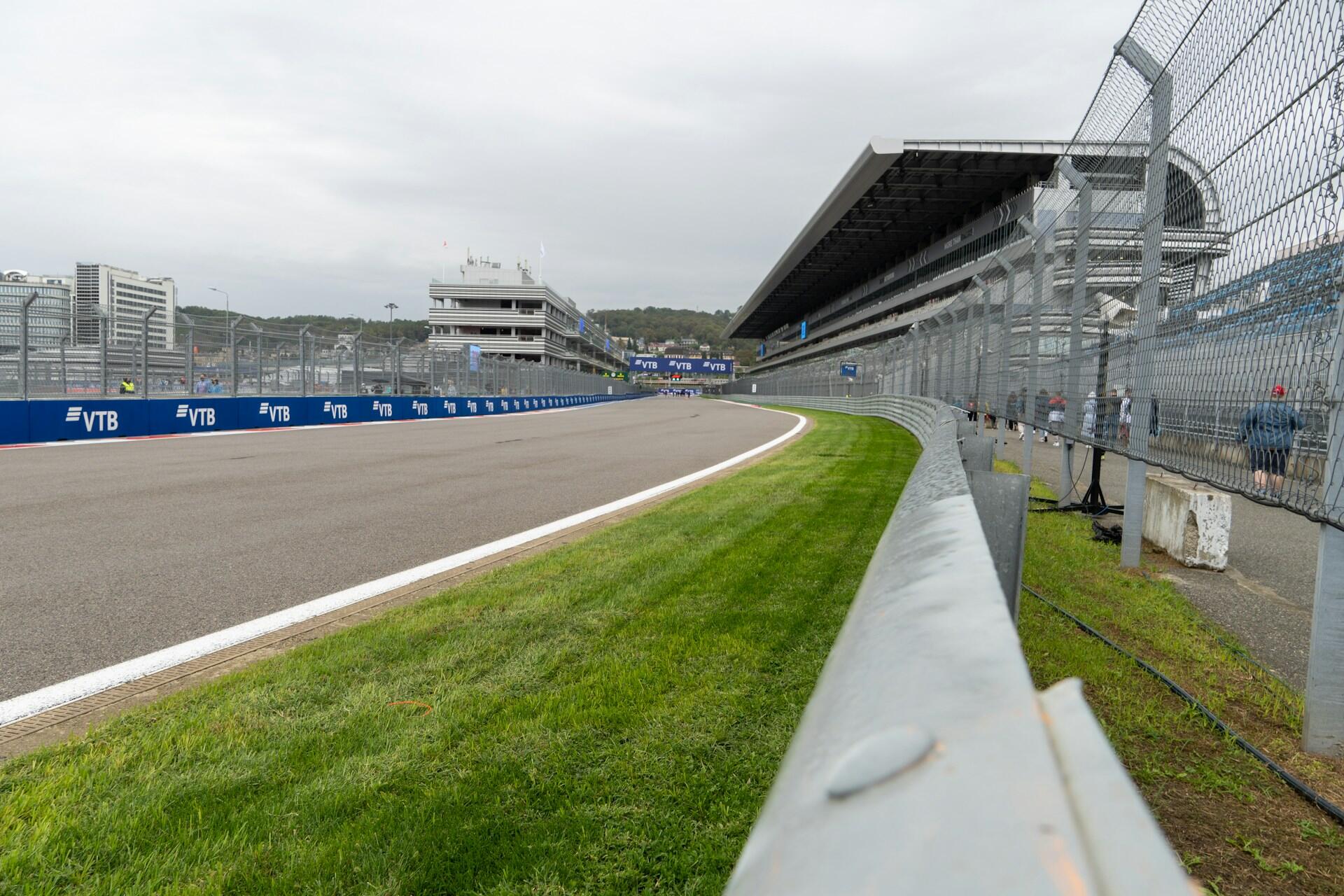 Race track with safety barriers, grandstands, and buildings under an overcast sky, with a few people visible near the fence.