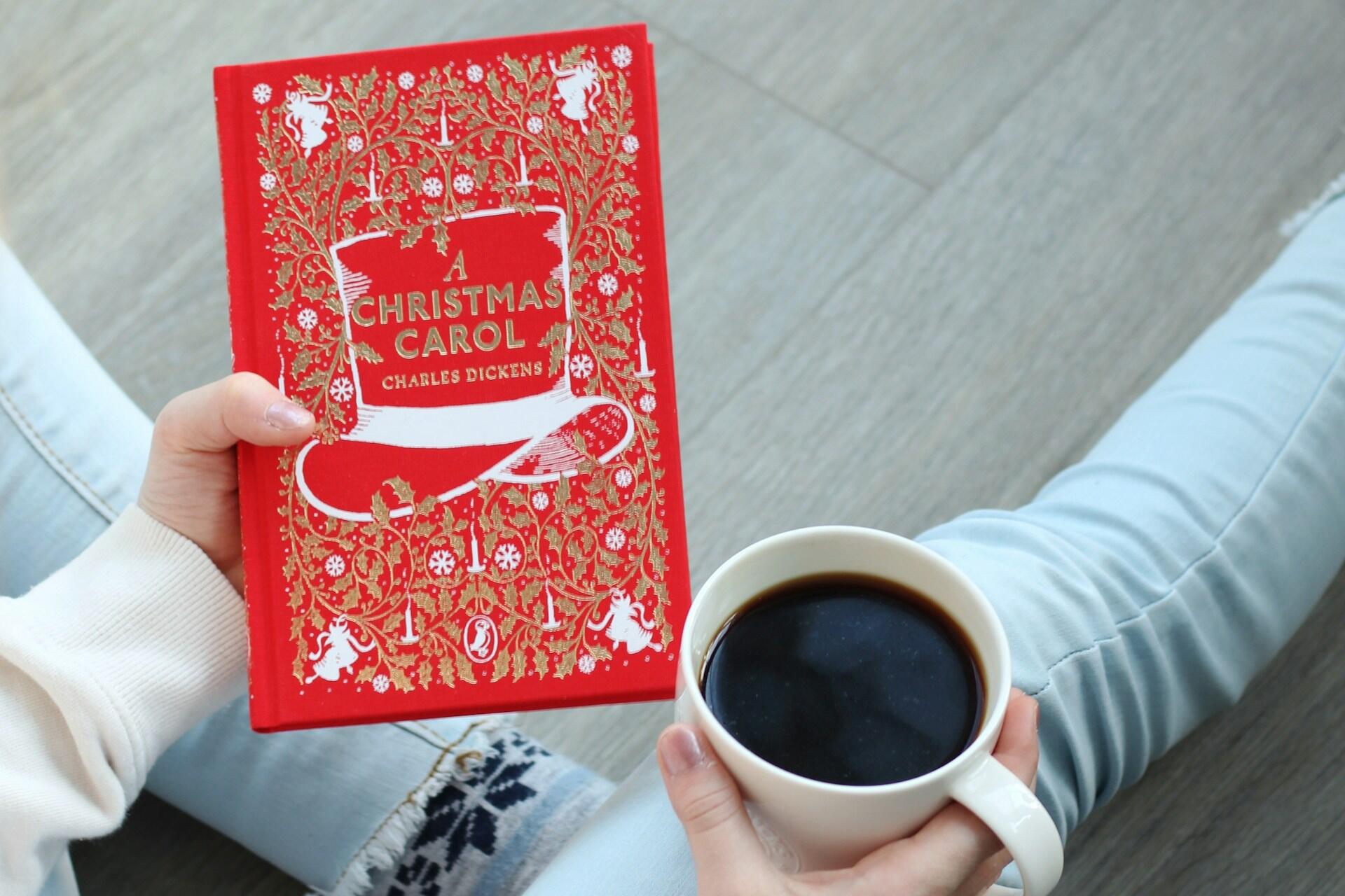A person holds a red book titled “A Christmas Carol” by Charles Dickens beside a white cup of black coffee.