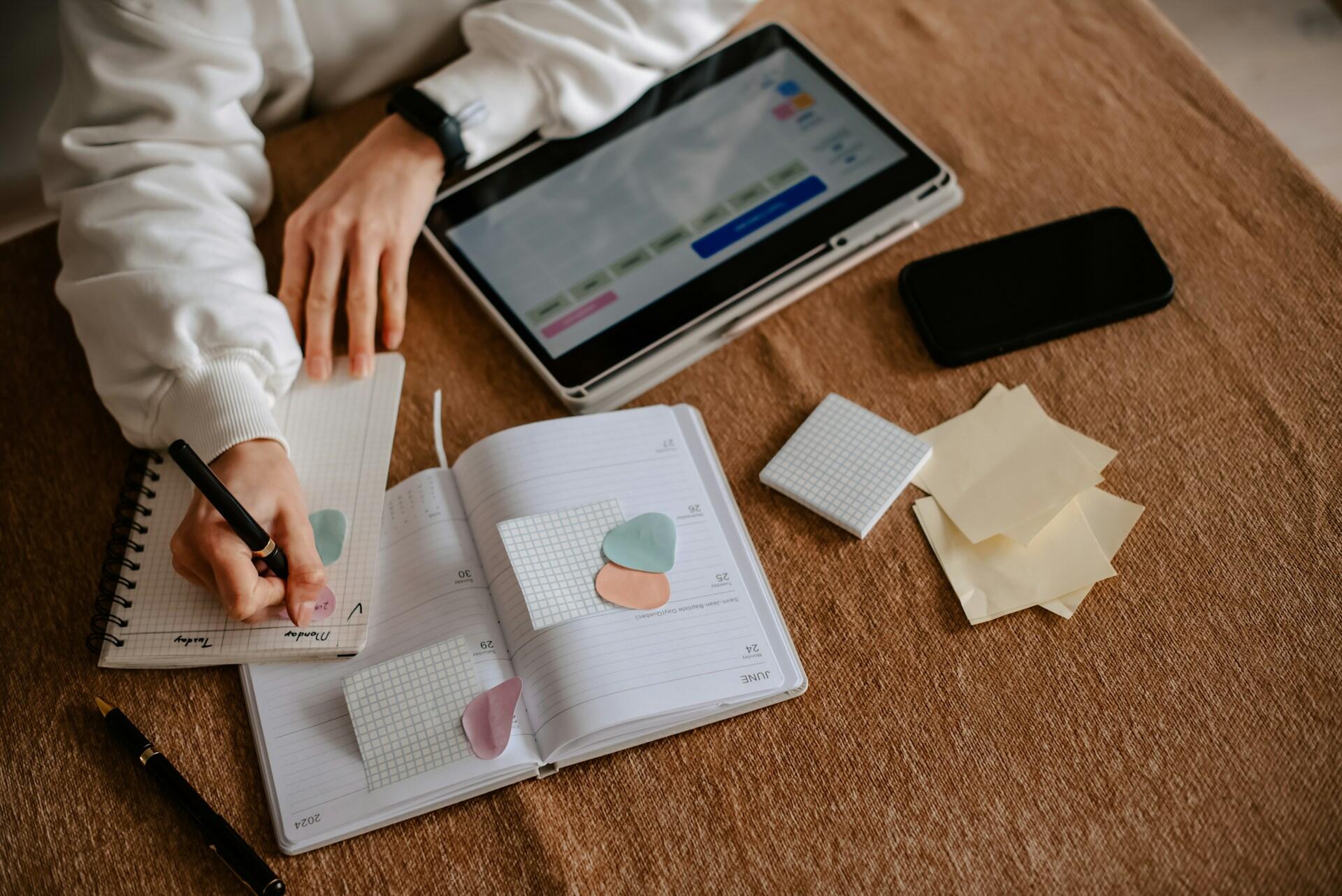 A person wearing a white shirt writes in a notebook, with a digital tablet open to a scheduler open in front of her.