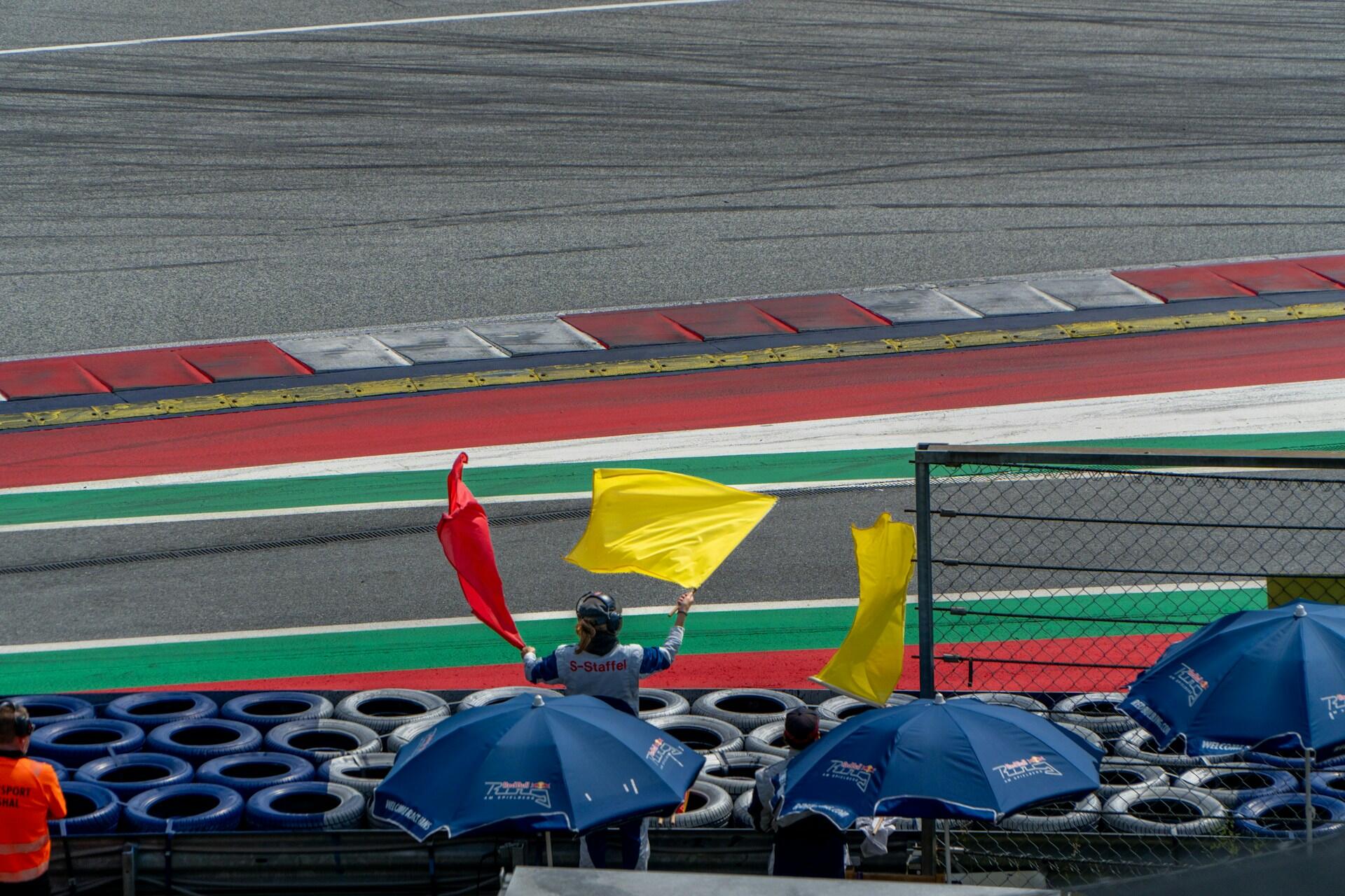 Race marshal waving red and yellow flags behind blue umbrellas near a race track with colored curbs and tire barriers.
