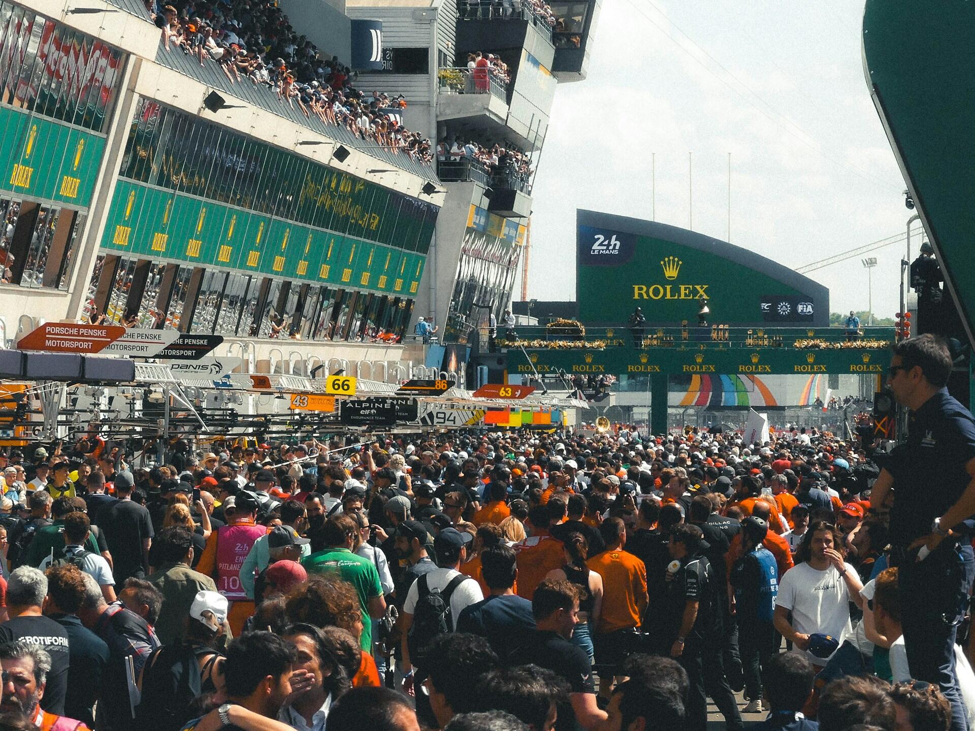Crowded pit lane at the 24 Hours of Le Mans race with teams, officials, and spectators under Rolex banners and race signage.