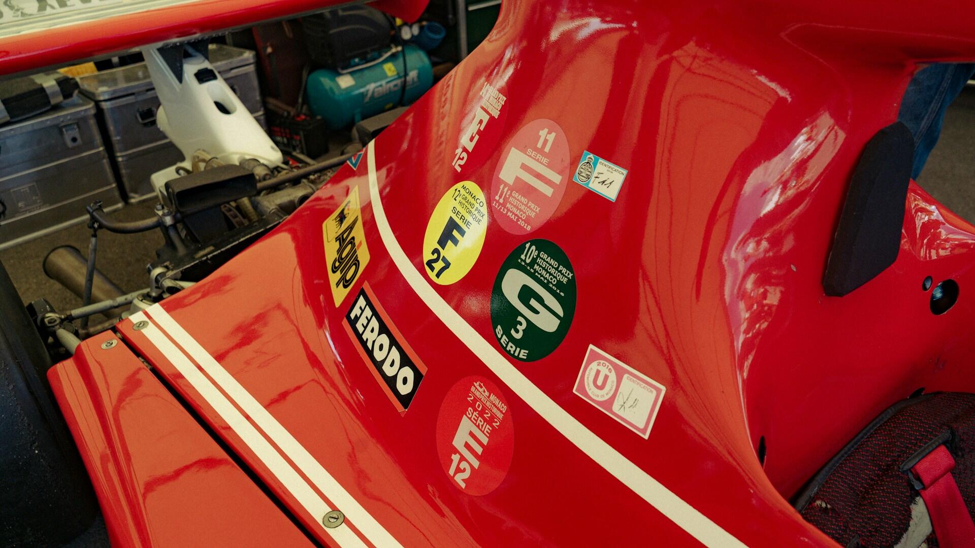 Close-up of a red race car body featuring various racing event stickers and sponsor logos near the cockpit area.