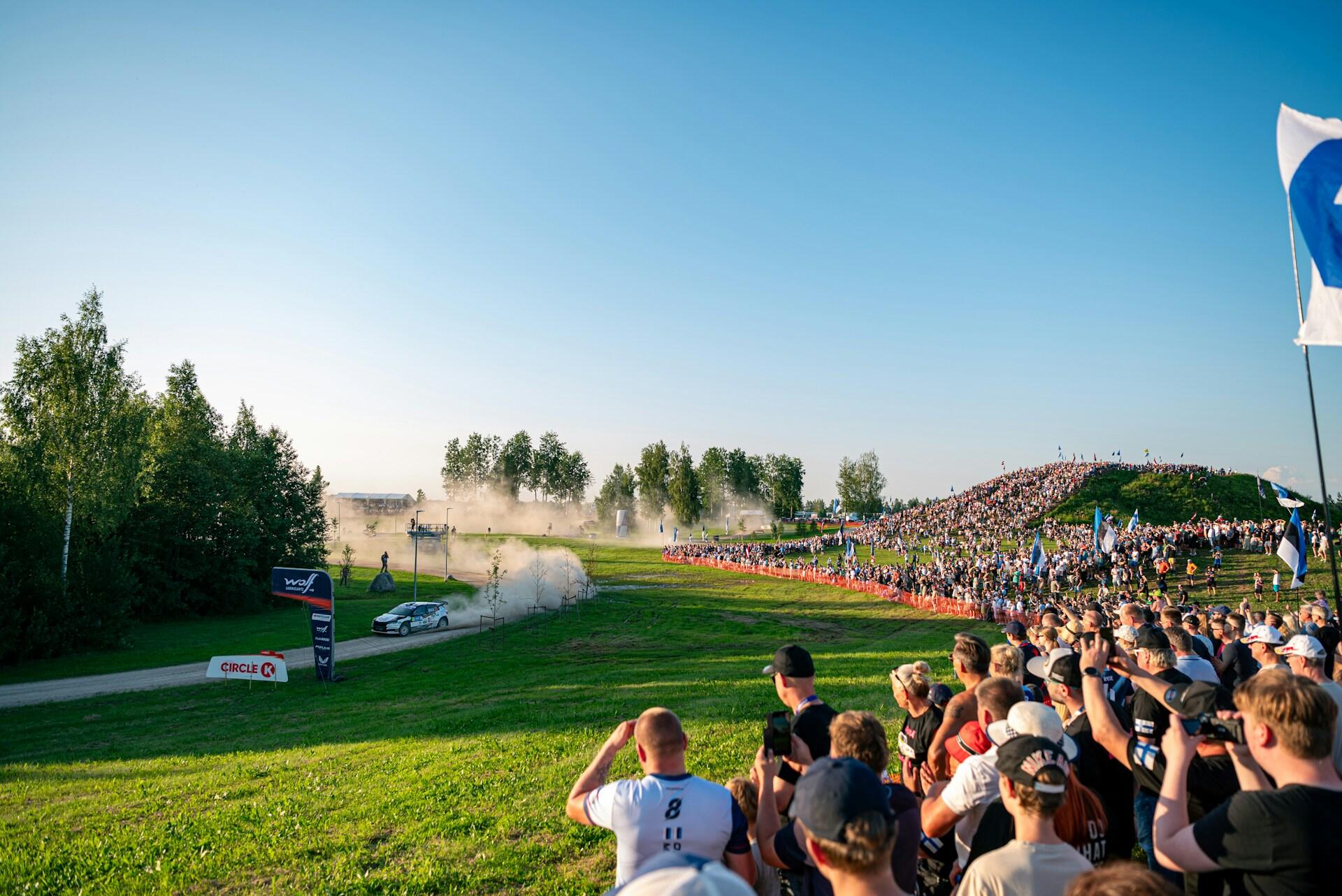Crowd watching a rally car speeding on a dirt track, kicking up dust, under a clear blue sky with trees and waving flags nearby.