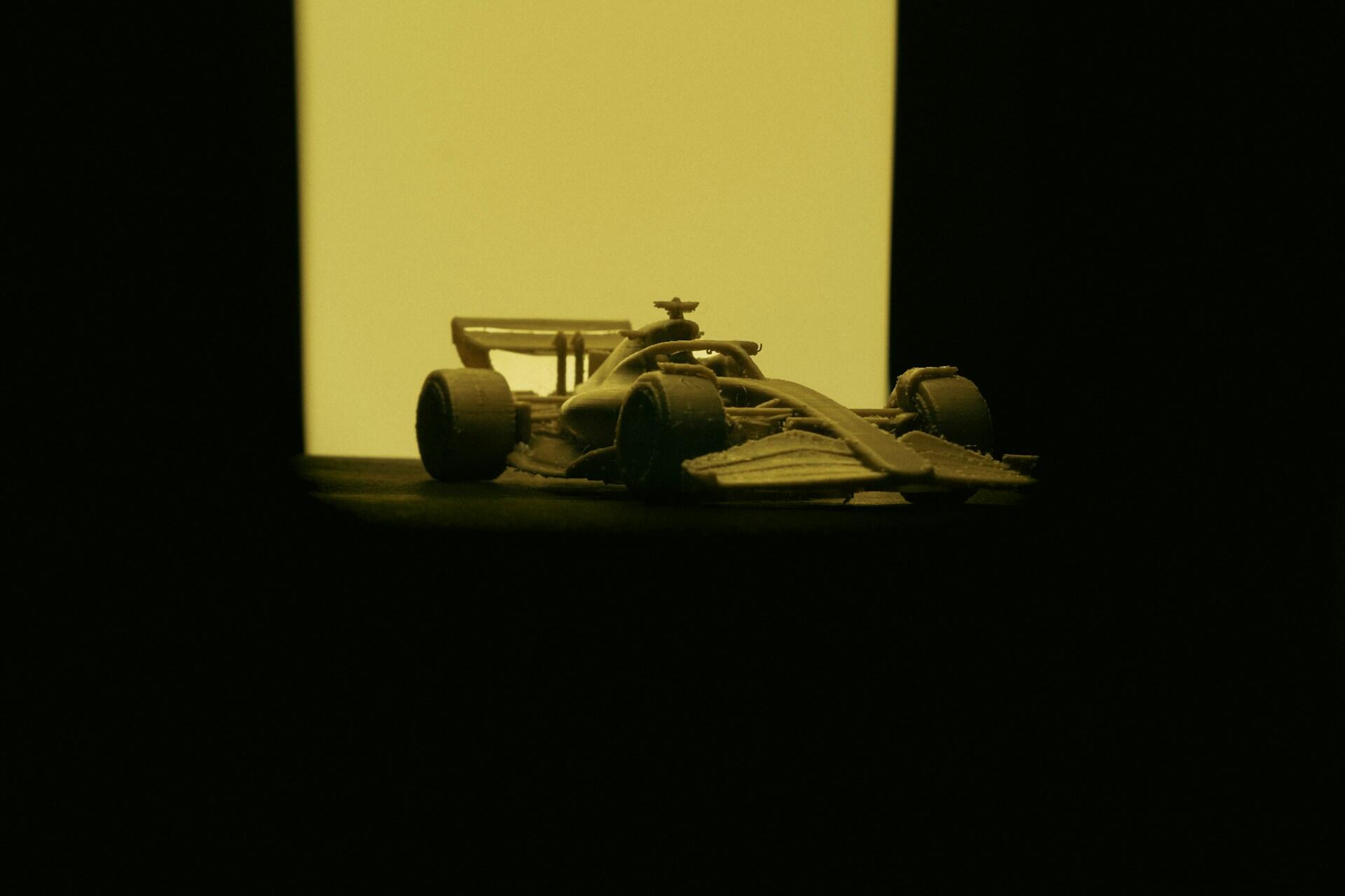 Miniature Formula 1 race car silhouette illuminated from behind by a yellow rectangular light in a dark setting.