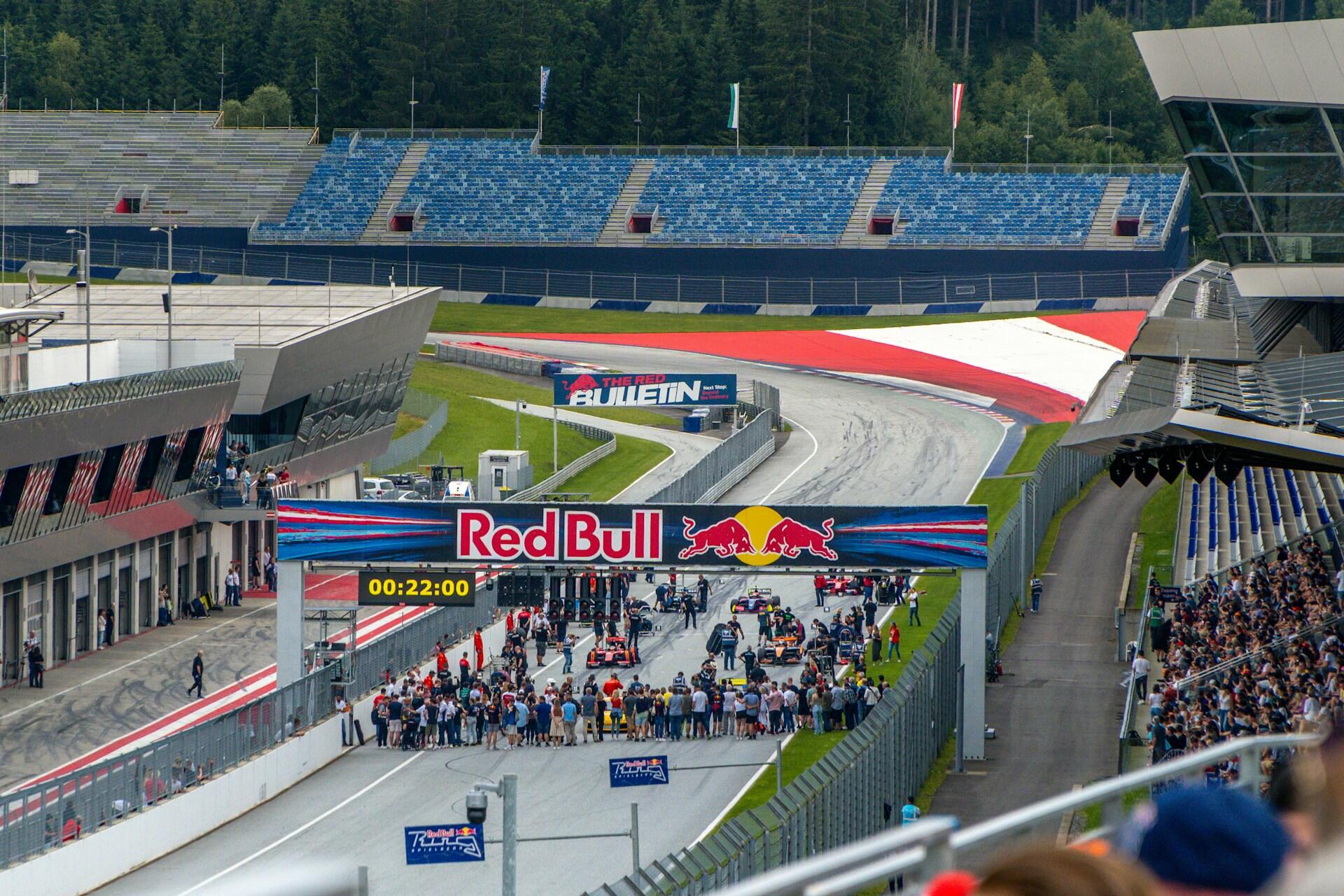 Crowd gathered around race cars on a Red Bull racetrack with empty grandstands and a forest in the background.