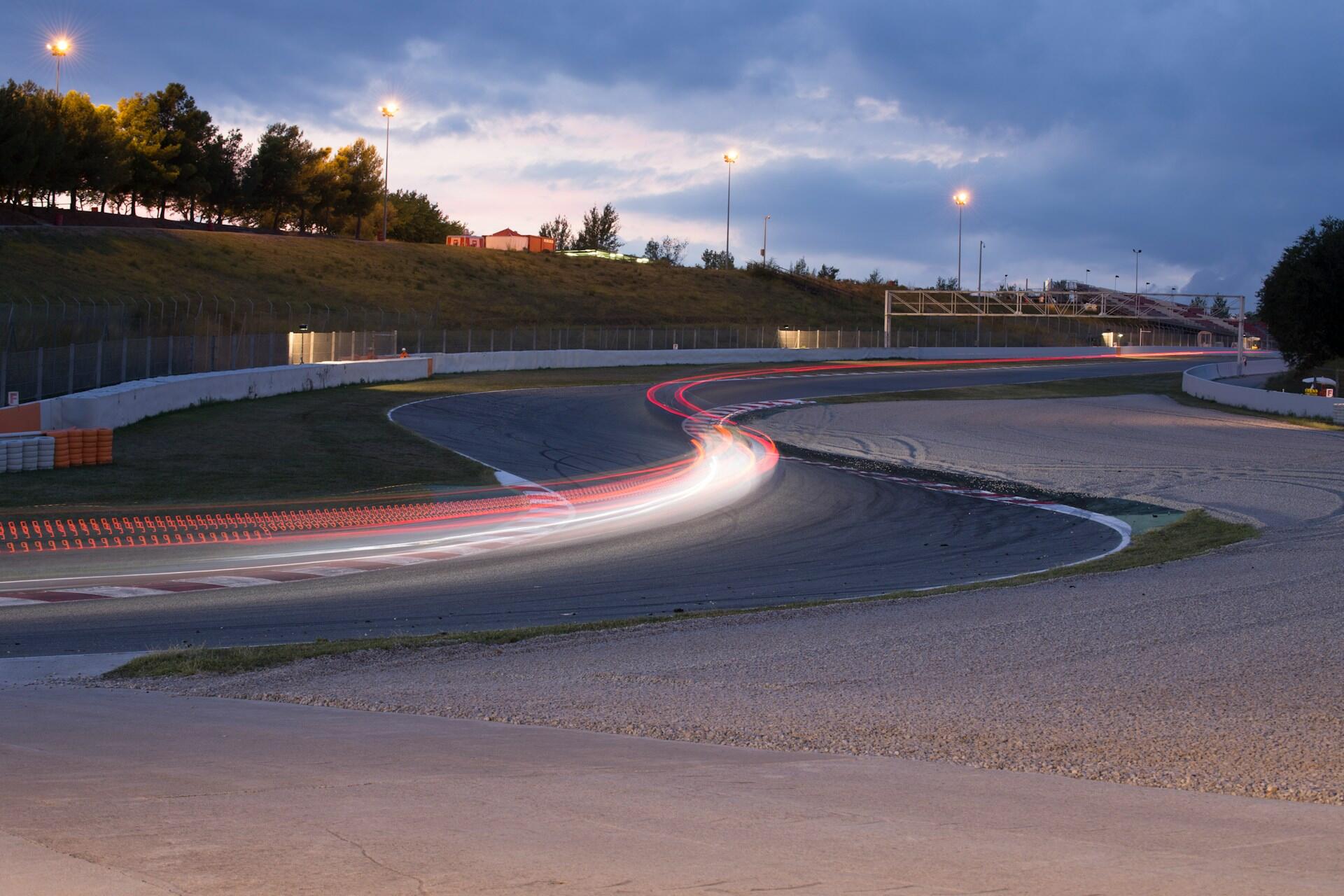 Race track curve at dusk with light trails from fast-moving vehicles and illuminated lampposts against a cloudy sky.
