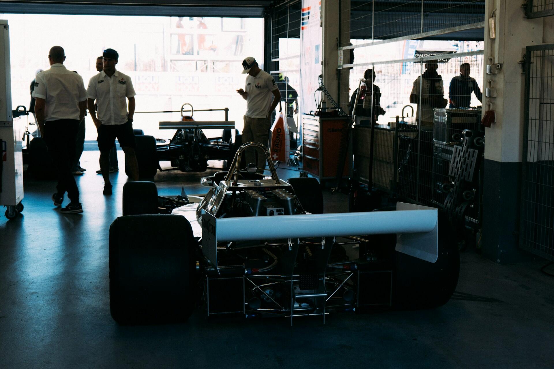 Formula-style race car in garage with people standing and walking around, equipment and tools nearby.