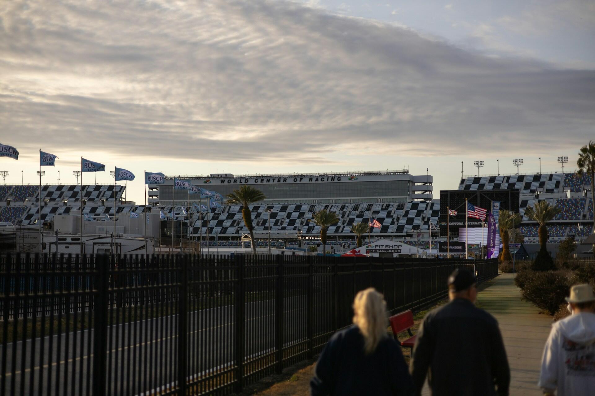 Three people walk toward the Daytona International Speedway grandstand with flags and palm trees under a cloudy sky.