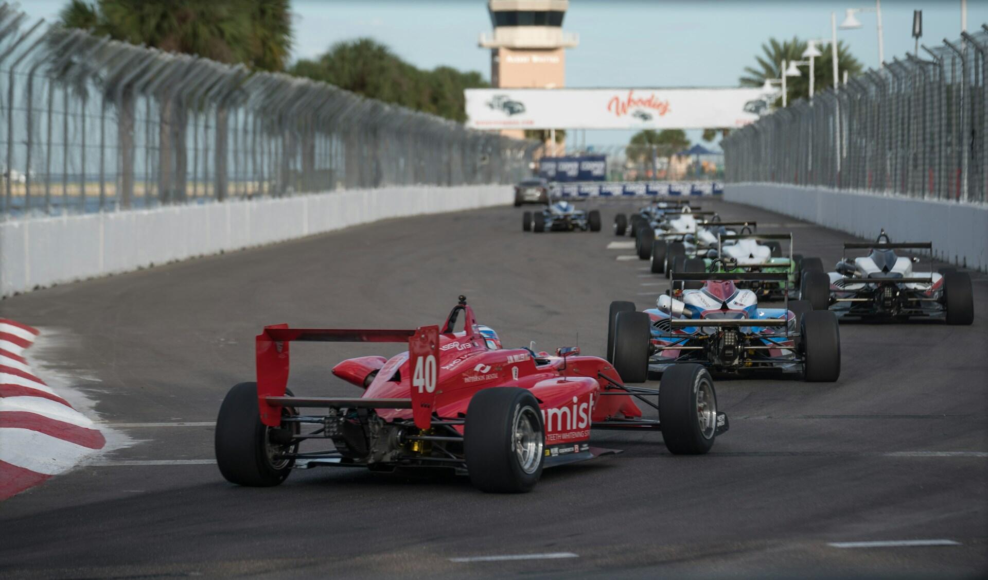Open-wheel race cars competing on a fenced asphalt track, navigating a curve with a control tower and banner in the background.
