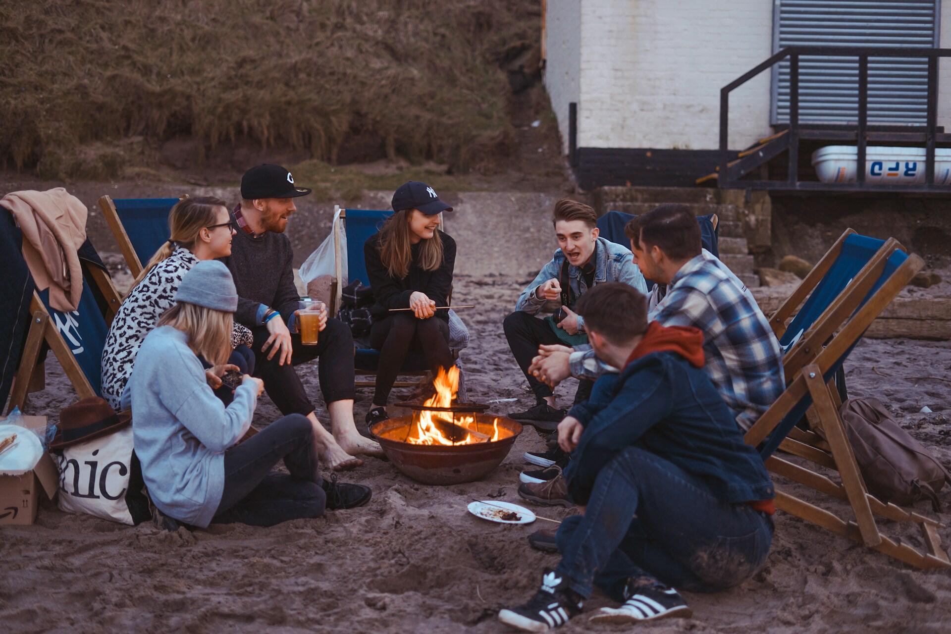 A group of friends in warm clothing sit around a campfire, sharing a laugh.