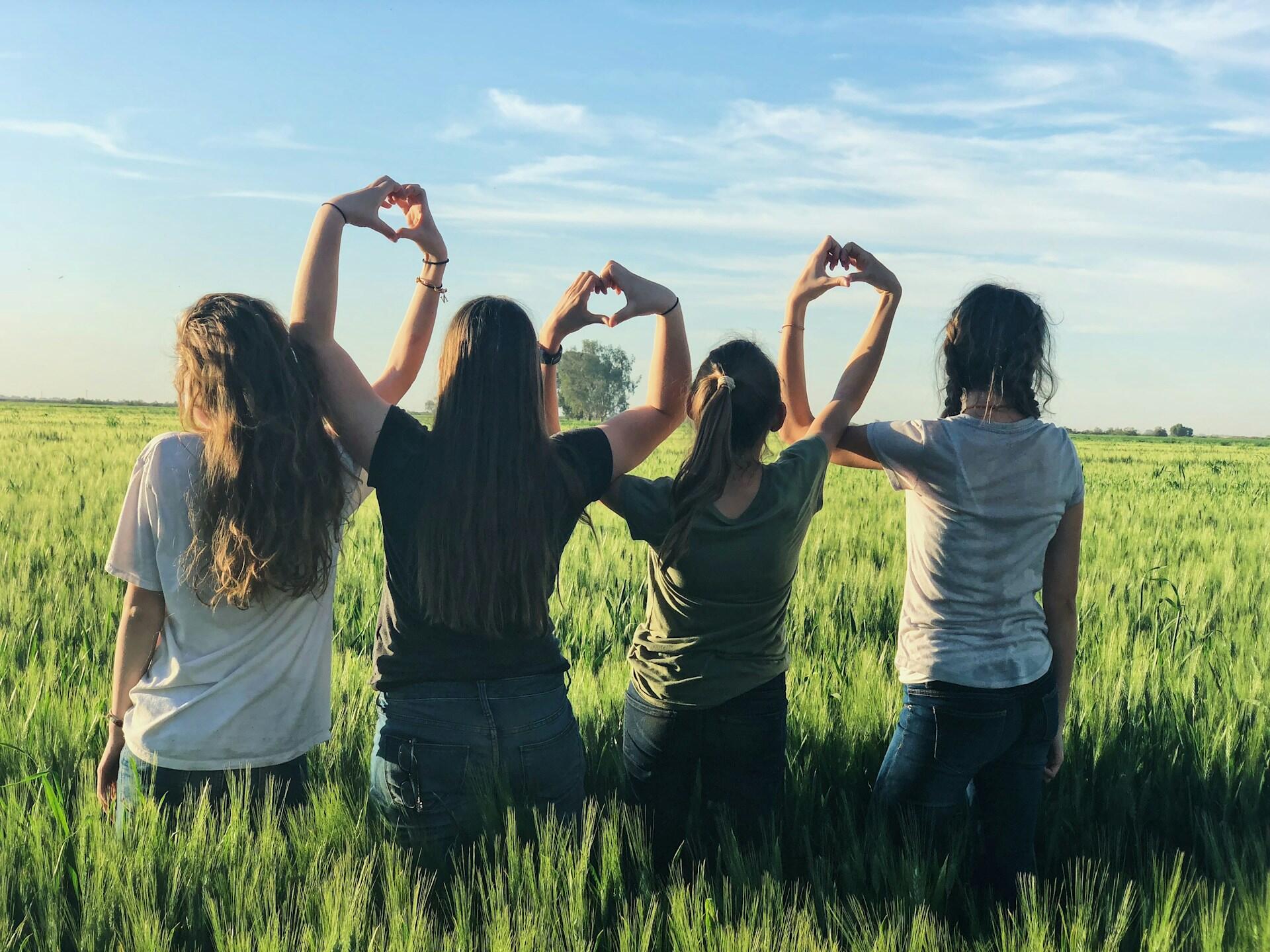 Four young women wearing casual clothing stand in a field holding their arms up with their interlocked hands forming heart shapes.