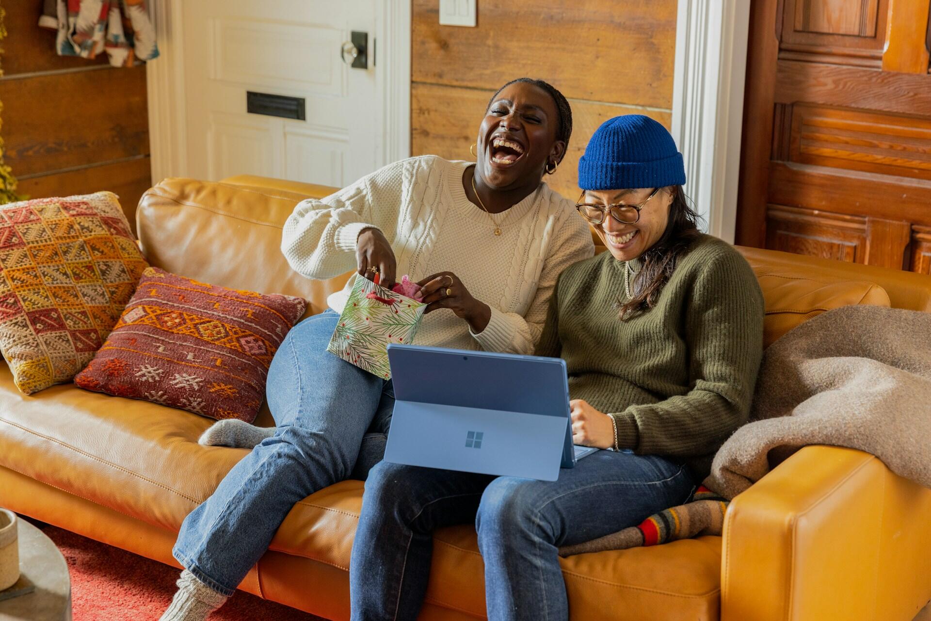 Two women sit on a couch laughing, while one holds a small gift bag and the other works a computer.