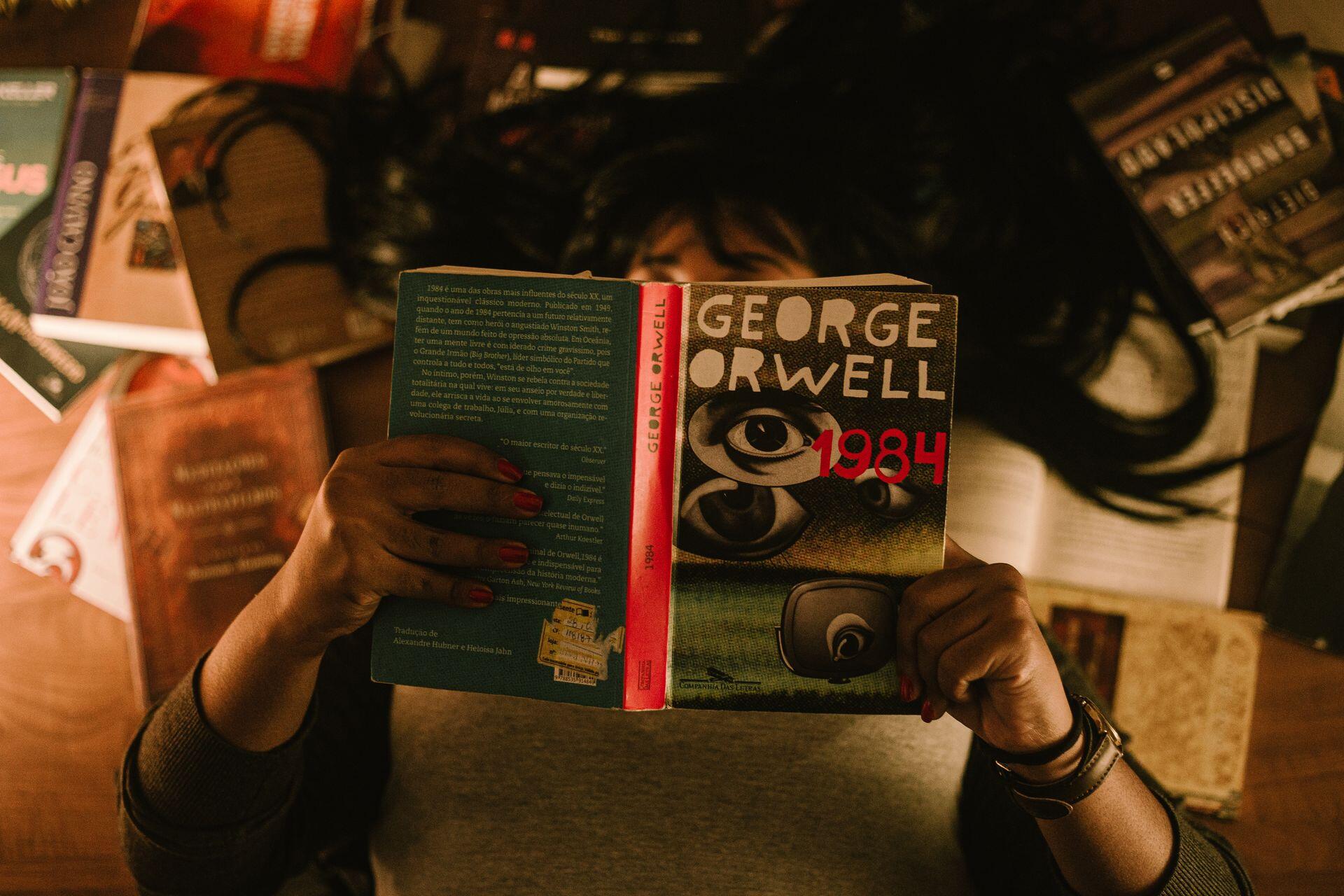 A person holds George Orwell’s book 1984 open in front of their face, surrounded by books and papers on a desk.