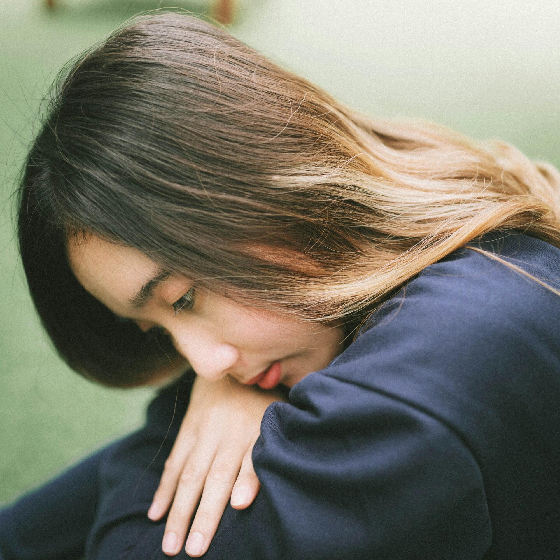 A young woman with long hair huddles with her face leaning on her knees, looking sad.