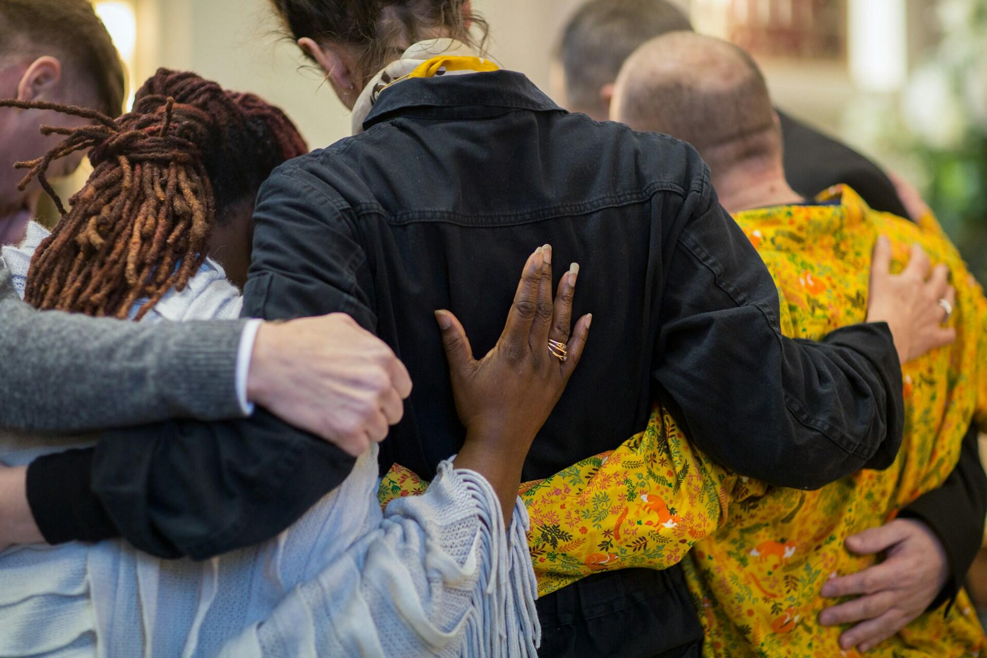 A group of people stand close together, with their arms around each other and their heads bowed. The mood is sad.