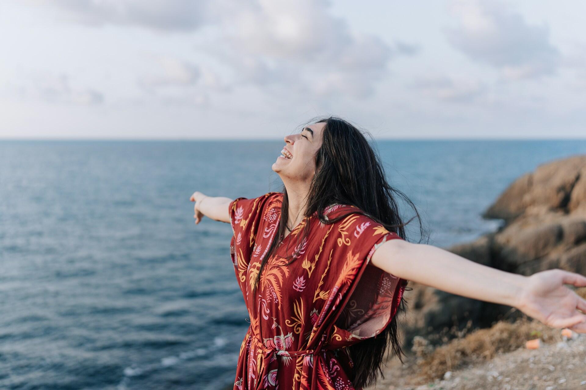 A woman wearing a comfortable orange garment stands on a beach on a sunny day with her arms spread wide, her head thrown back and smiling wide.