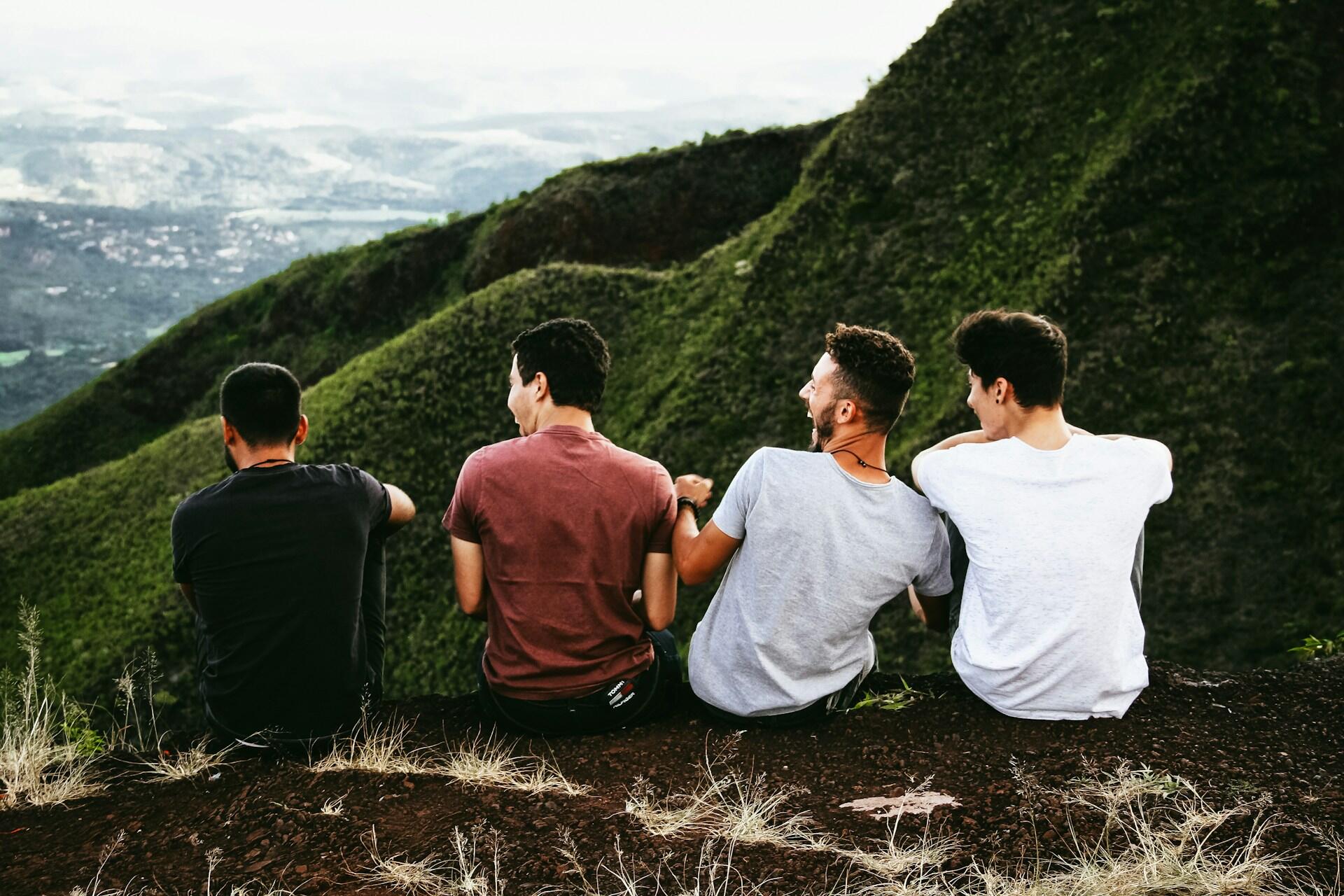 Four young men sit together atop a mountain, laughing and gazing out at the greenery of other mountain faces.