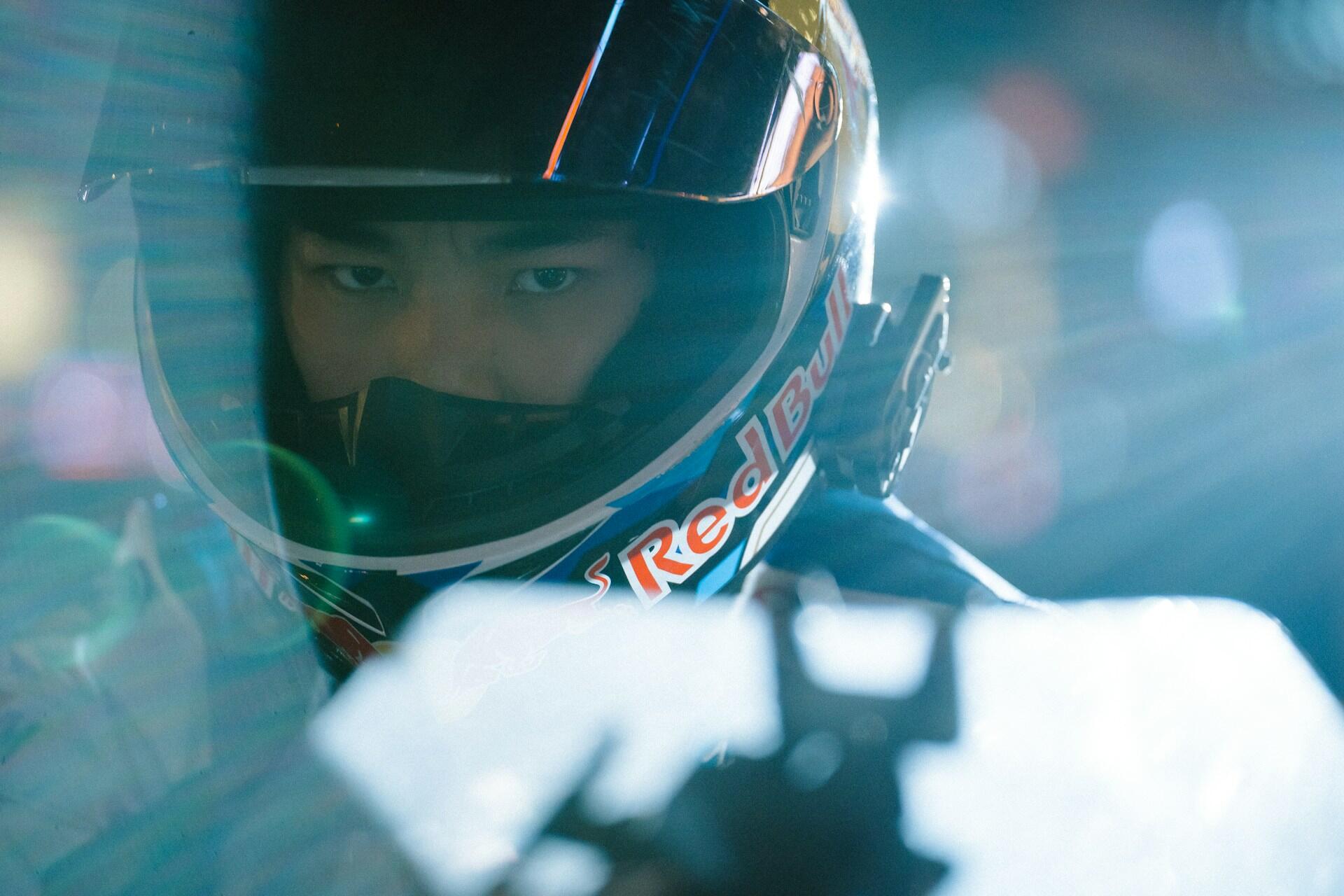 Close-up of a race car driver wearing a Red Bull helmet, focused and ready to compete under bright lights.