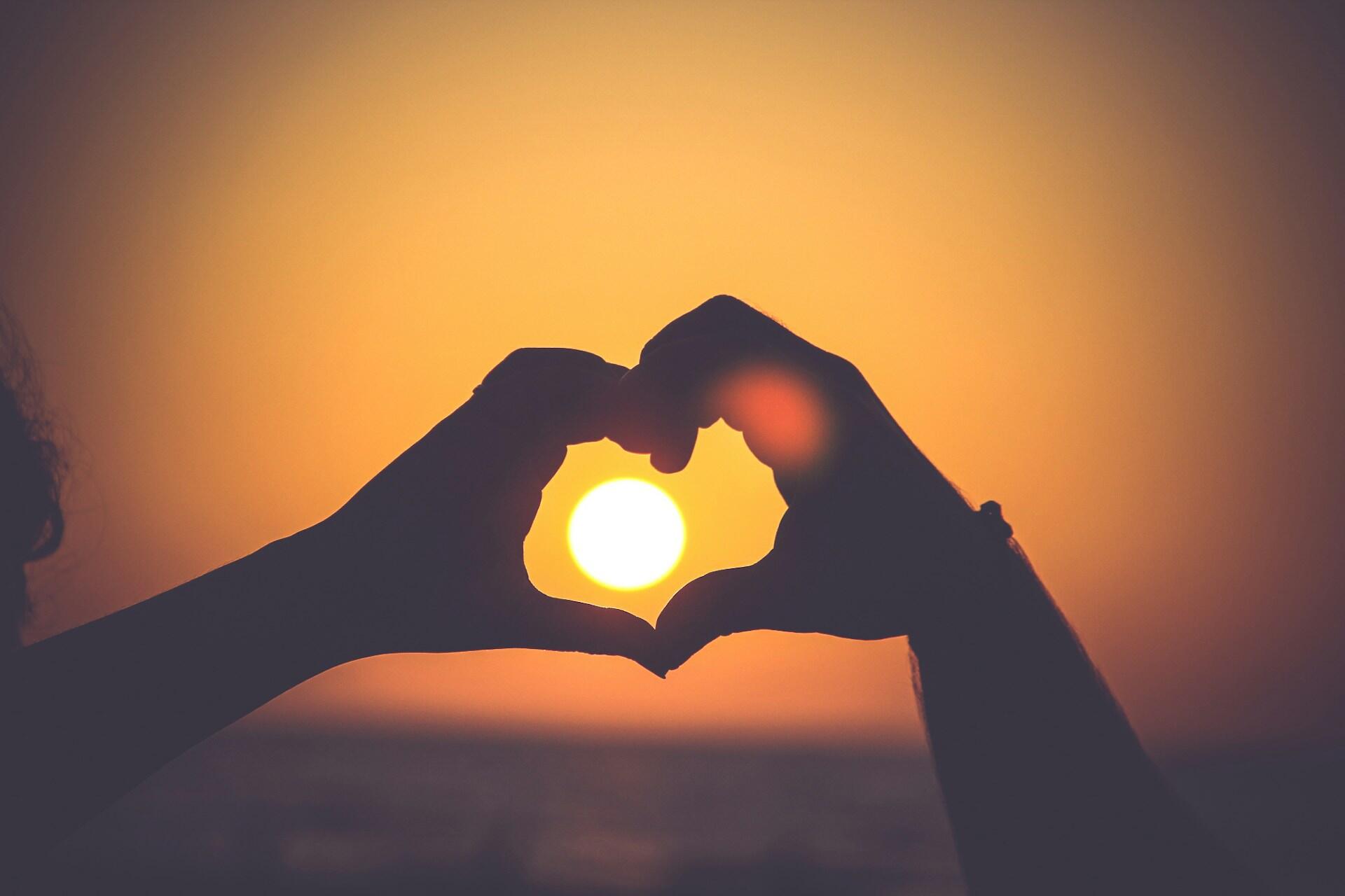 Two hands create a heart silhouette, illuminated by a glowing sun in the background, representing love and connection.