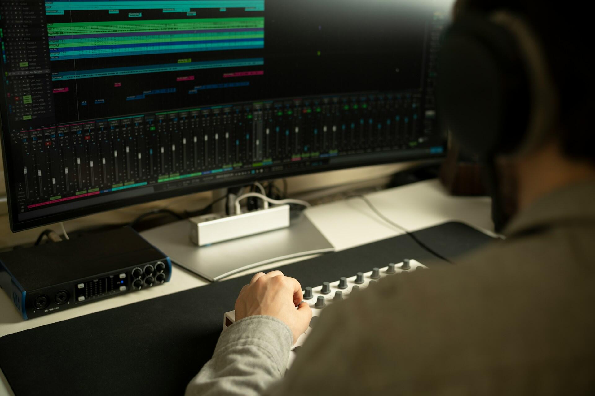 mastering engineer sitting at a computer and using knobs to adjust tracks displayed on the DAW