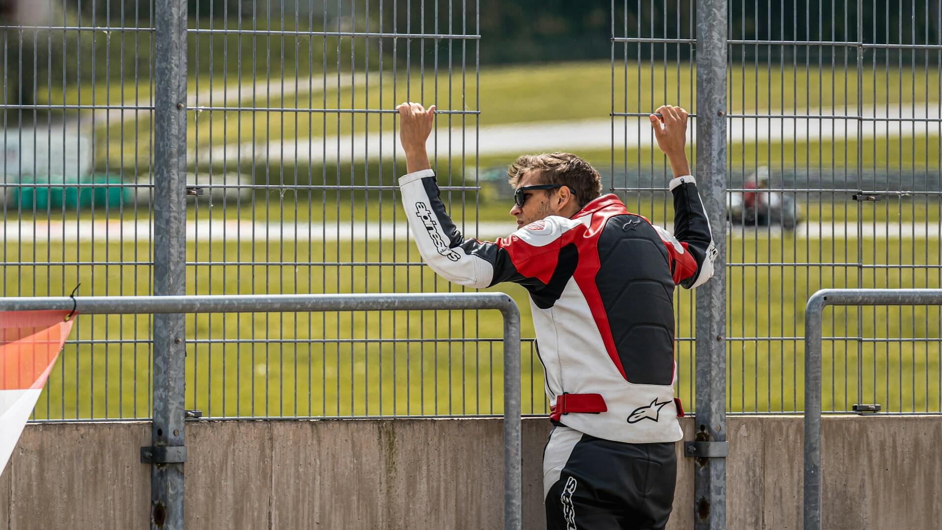 Man in red, white, and black motorcycle gear holding onto a metal fence, with a racetrack and blurred vehicle in the background.