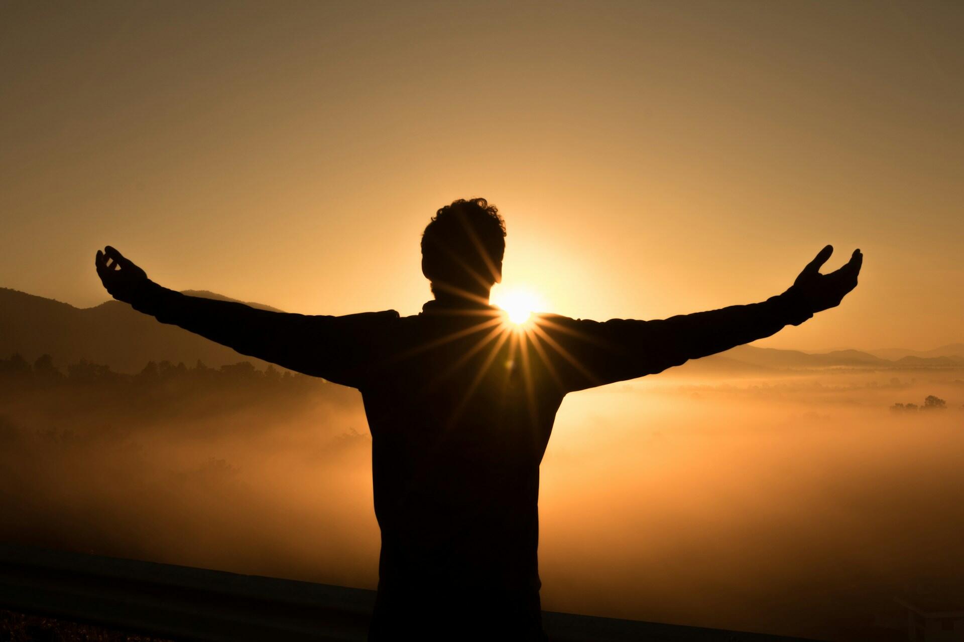 The silhouette of a person on a hilltop with arms stretched wide and the setting sun giving the image a golden glow.