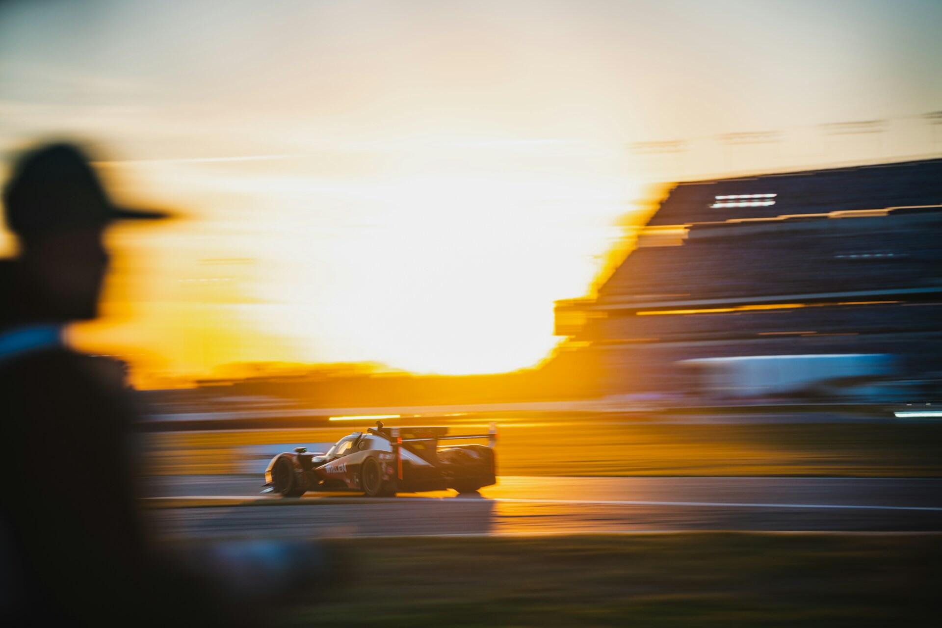 Race car speeding on a track at sunset with a blurred person in the foreground and grandstands in the background.