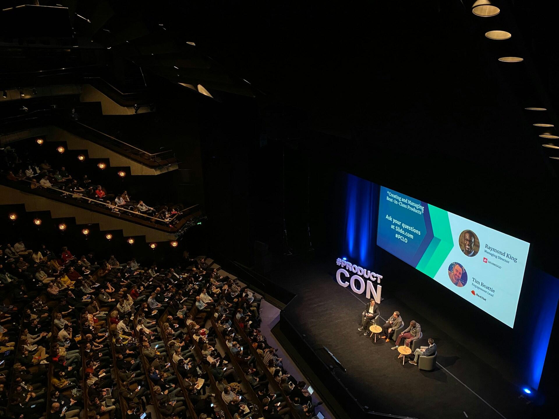 an auditorium full of audience members looking at a pit stage with four guest speakers sitting in chairs and talking. A large LED screen behind them explains their names and titles and a physical sign indicates the event is happening at ProductCon