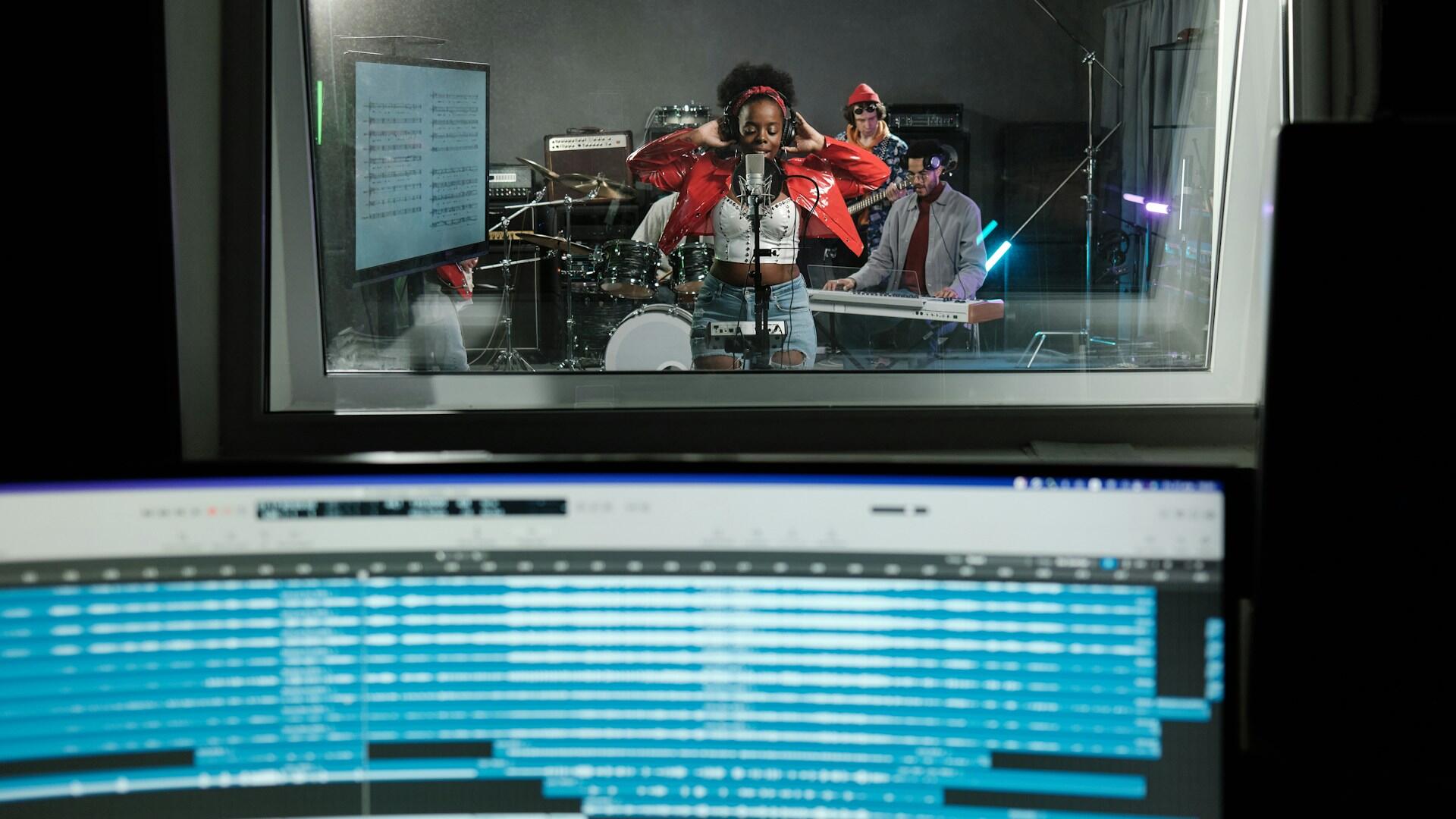 a band in a recording studio playing instruments and singing while a recording engineer in the booth monitors the recording
