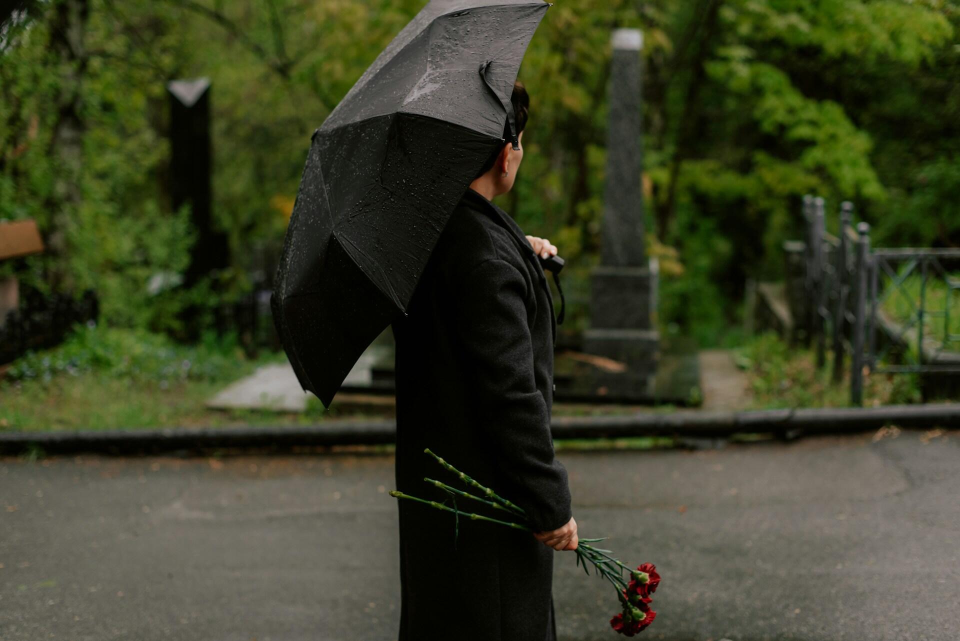 A person wearing black carries an open black umbrella and three red carnations while standing in a graveyard. The mood is sad and wistful.