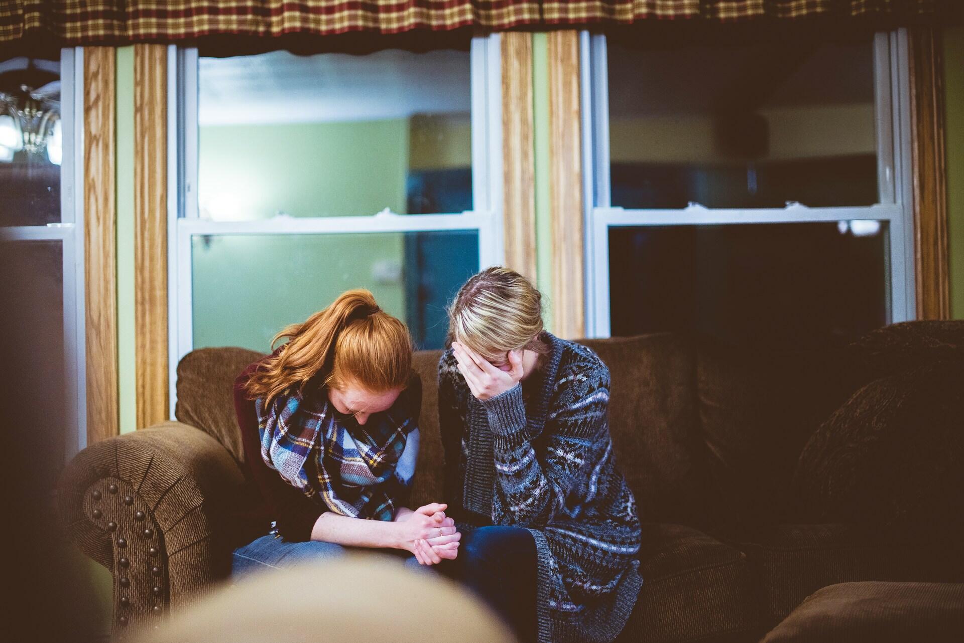 Two women sit on a couch, clutching each other's hands and hiding their faces. The mood is sad and tragic.