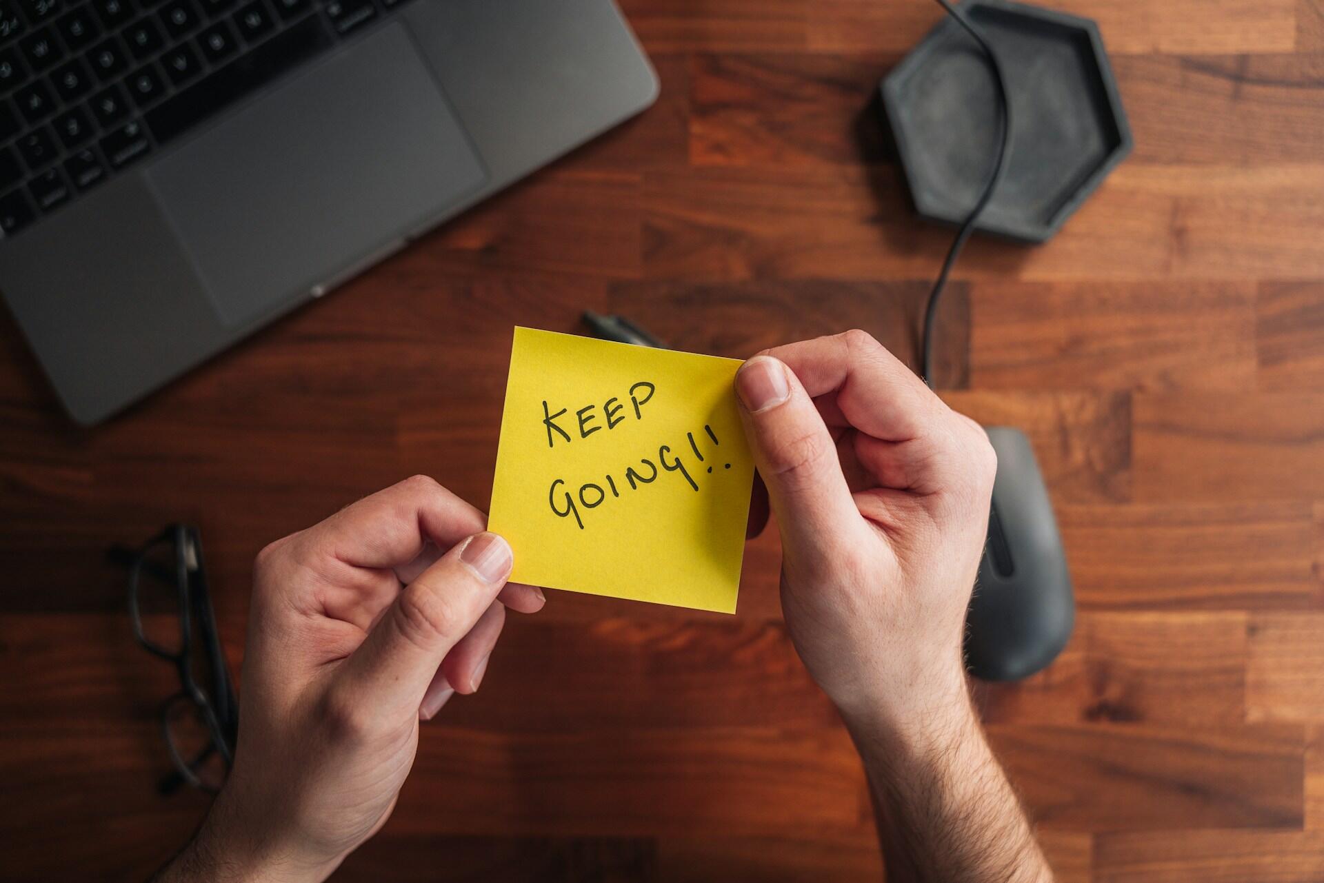 A person holding a yellow Post-It note with the words 'keep going' written on it.