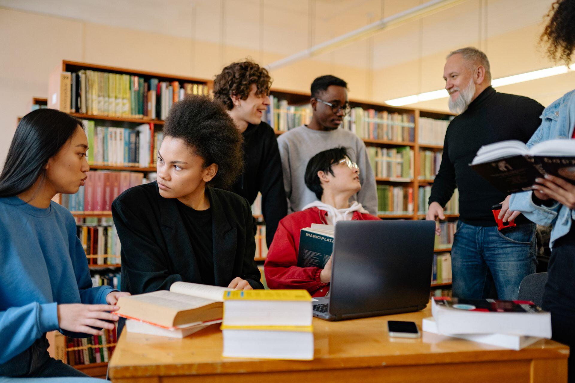 Diverse students and an older instructor gather around books and a laptop at a library table, discussing their studies.