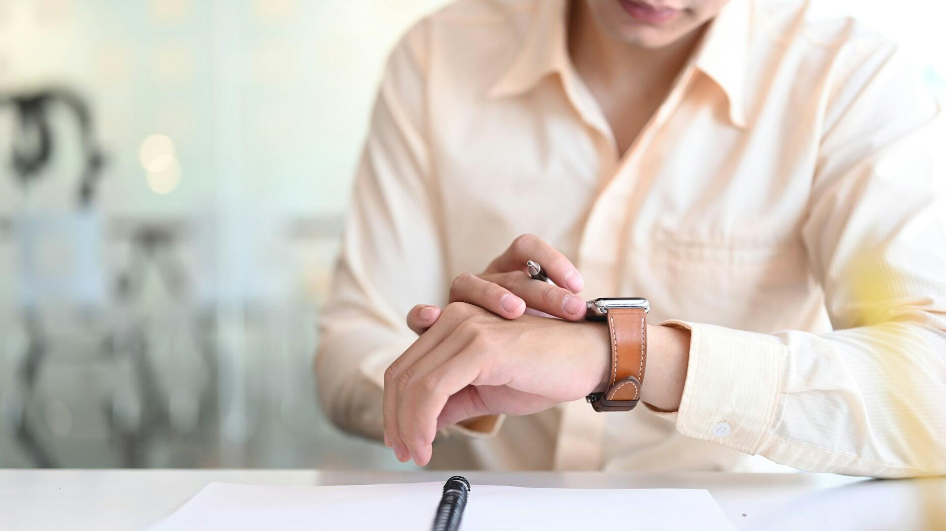 A person wearing a light-coloured shirt consults the watch he wears on his wrist.