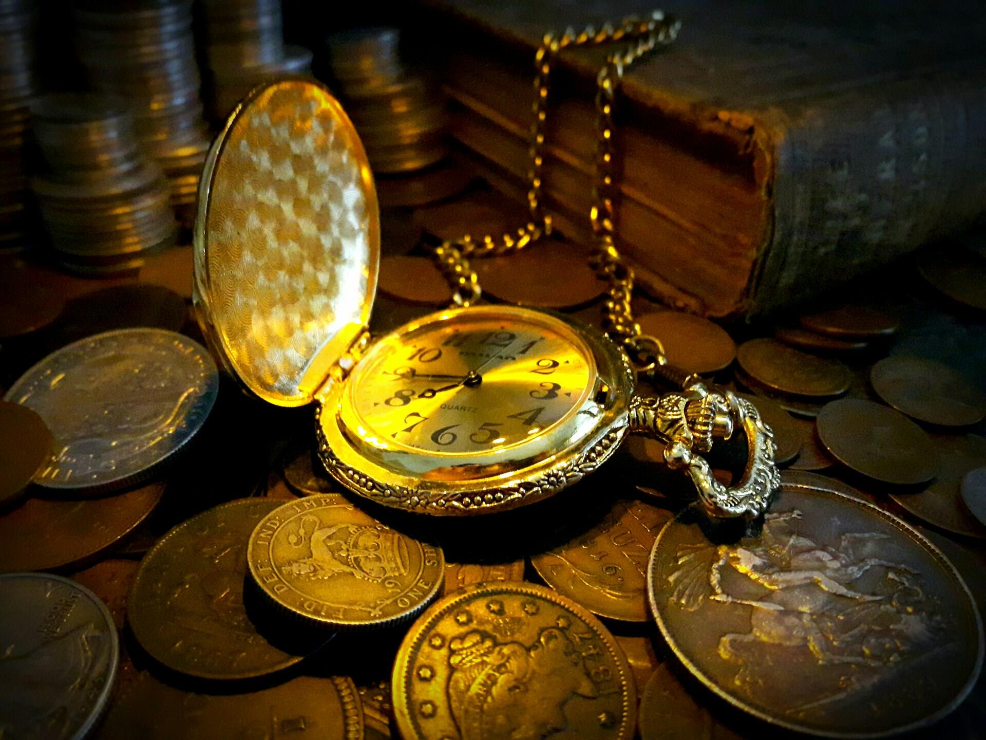 A golden pocket watch sits open on a pile of golden coins.