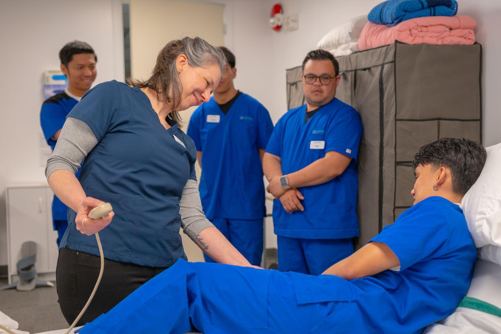 Junior nurses stood in background while senior nurse smiles and looks down at nurse lying on hospital bed