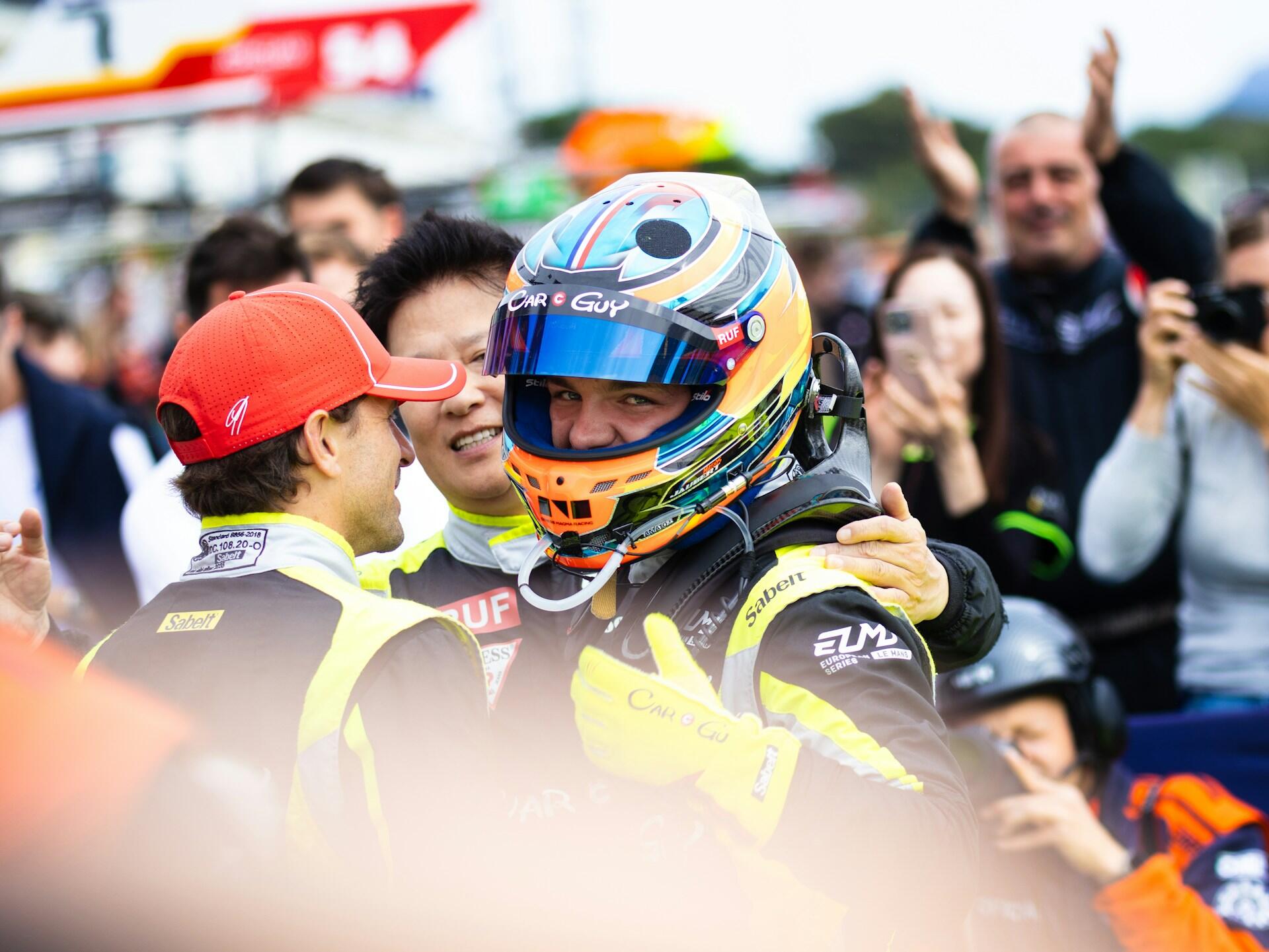 Race car driver in helmet celebrating with team and crowd applauding in the background at a racing event.