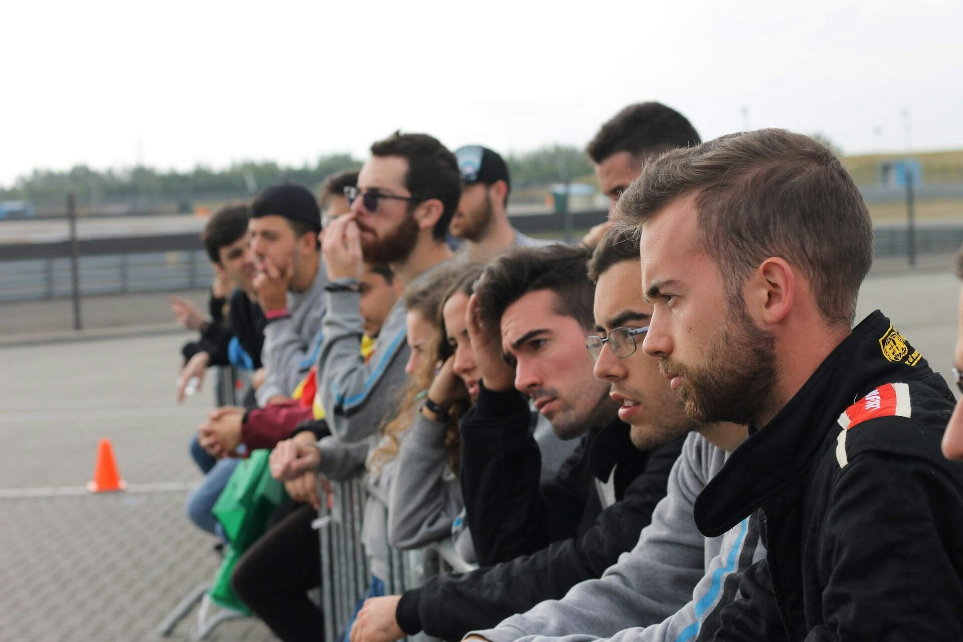 Group of young people closely watching an outdoor event behind a metal barrier, showing focused and contemplative expressions.