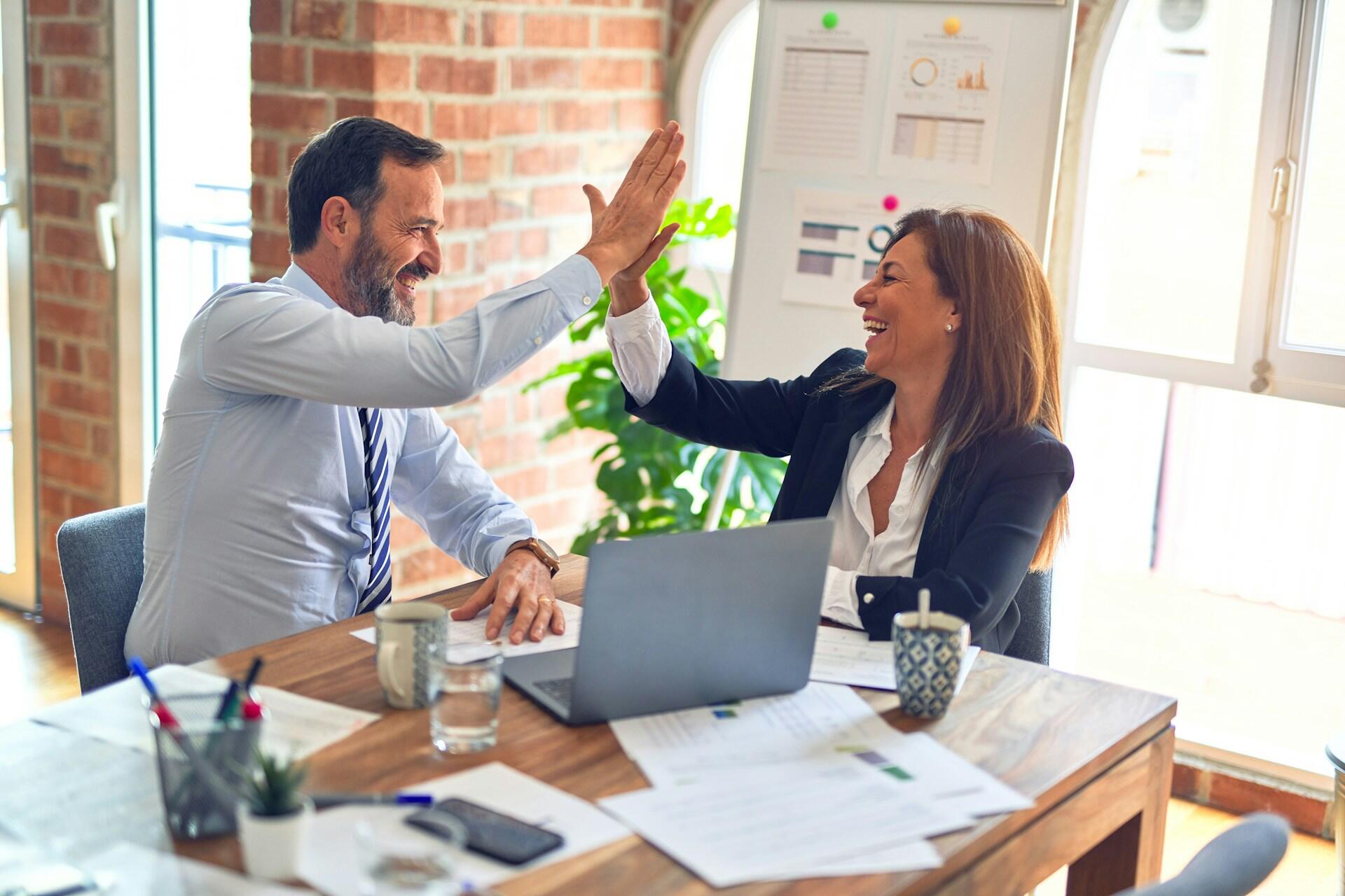 Two people wearing business attire sit at a table with a computer, looking happy and high-fiving each other.