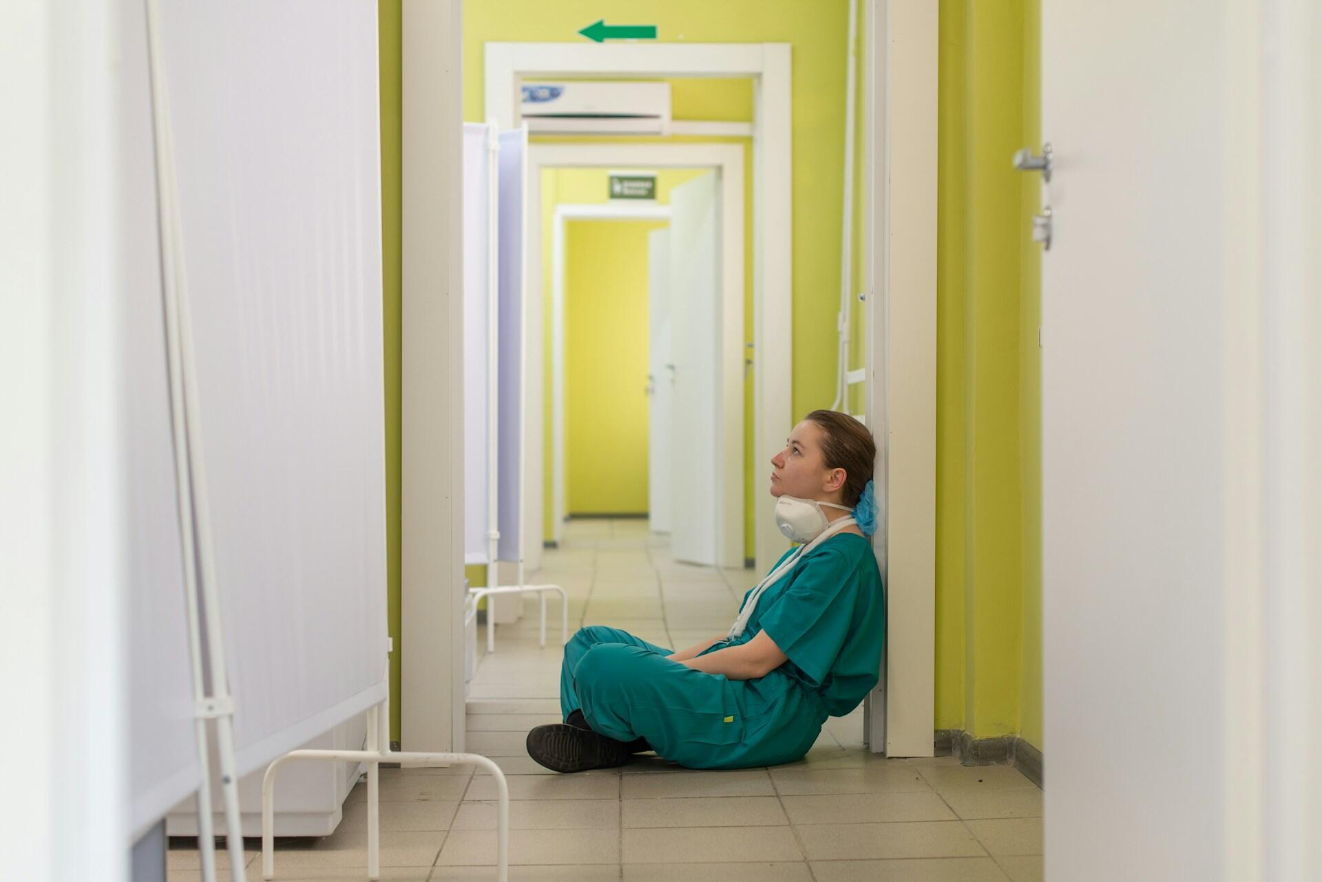 A person wearing green scrubs and a pulled-down face mask sits crosslegged in a yellow hall, looking despondent.