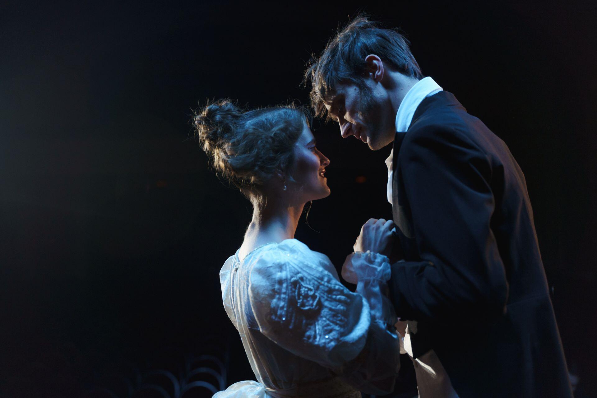 A man in a dark suit leans toward a seated woman in a light blue top as she looks up at him and holds his hands.