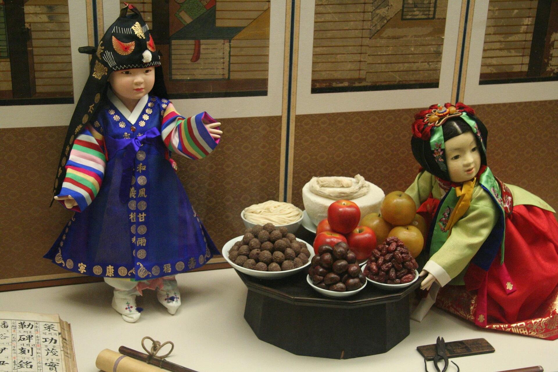 Two dolls representing a Korean boy and girl, model traditional clothing style while posed around a tray of tasty foods.
