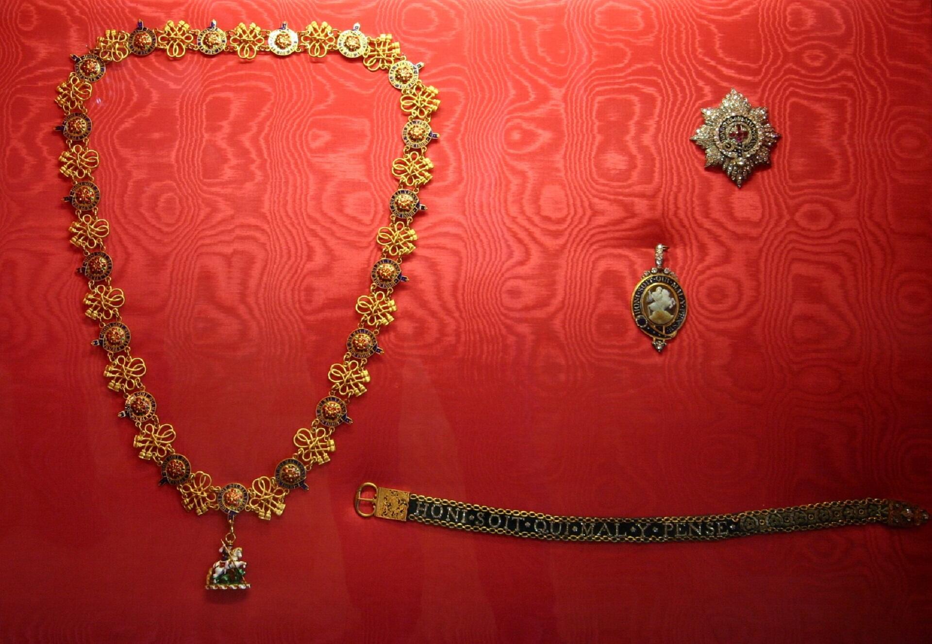 Close-up of a chivalry insignia, featuring a gold chain and pins, set against a red cushion, on display, highlighting their craftsmanship and artistic details.