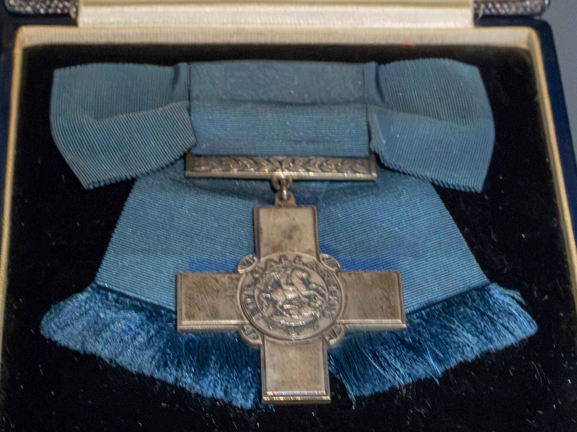 A silver cross with a light blue ribbon resting on a blue velvet background in a dark blue case.
