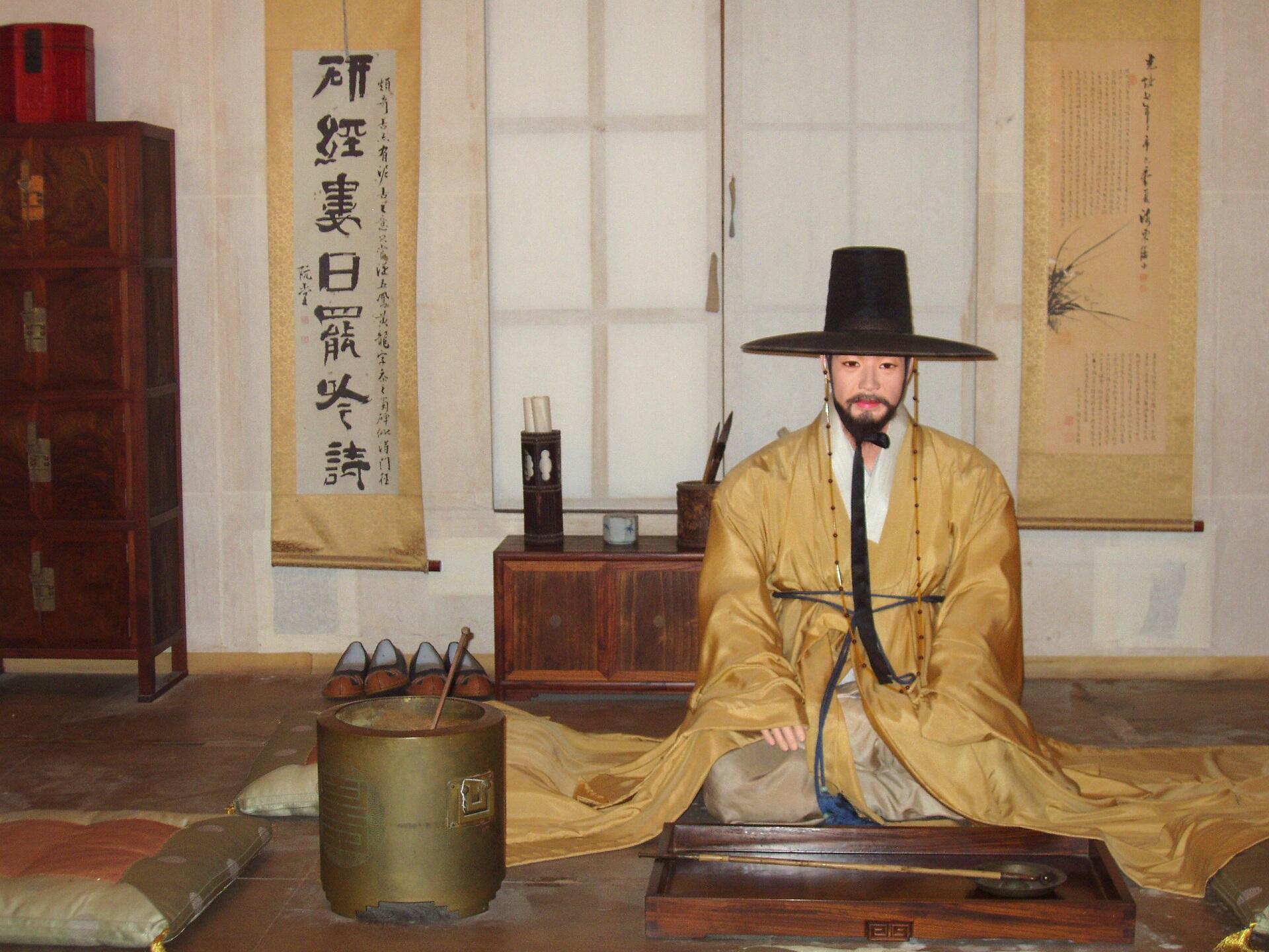 A man kneels on the floor in front of a window, wearing ancient Korean garb with a period hat, appearing solemn.
