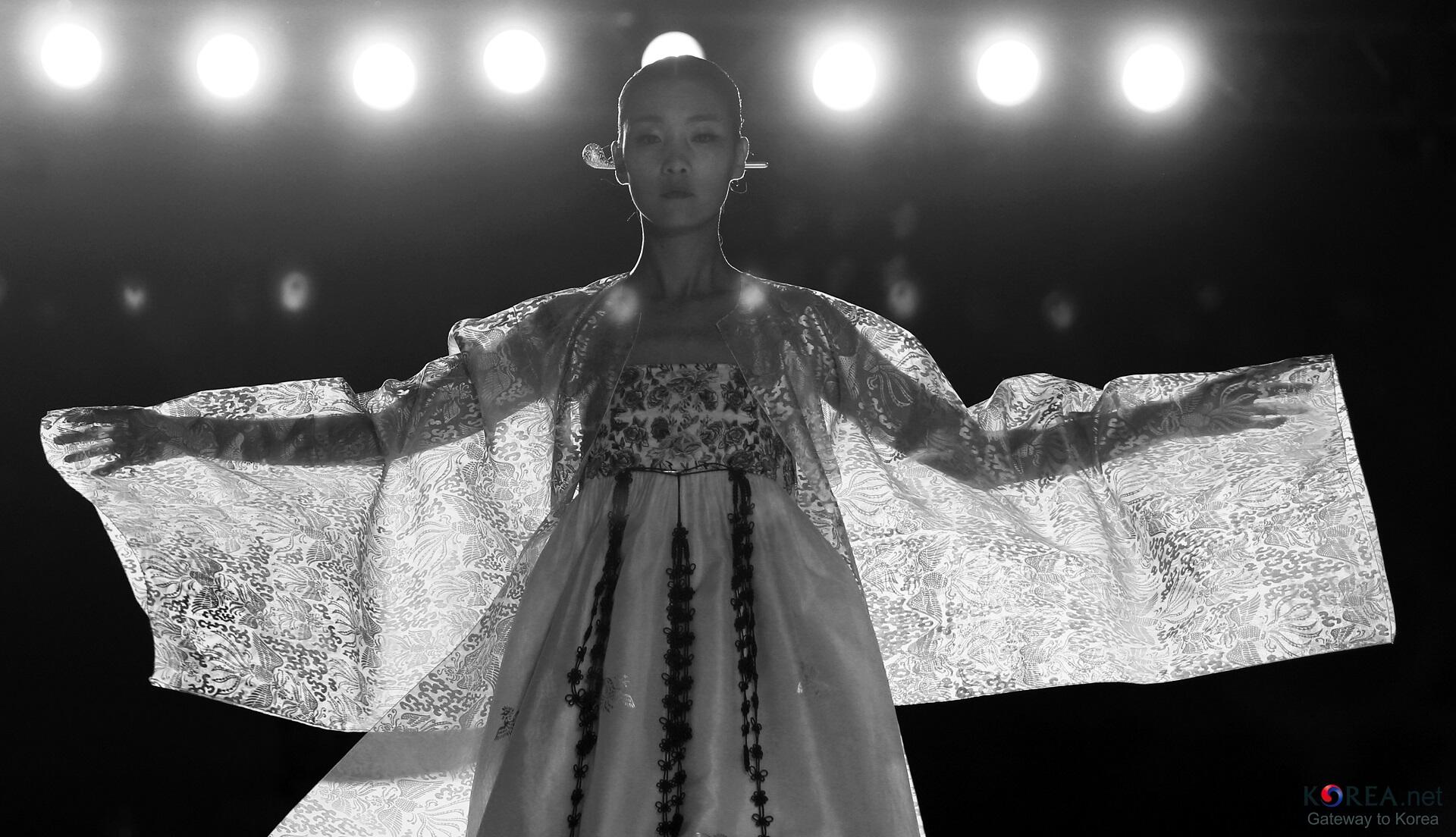 A young woman with her hair in a bun models a gossamer hanbok-style outfit on a fashion show runway.