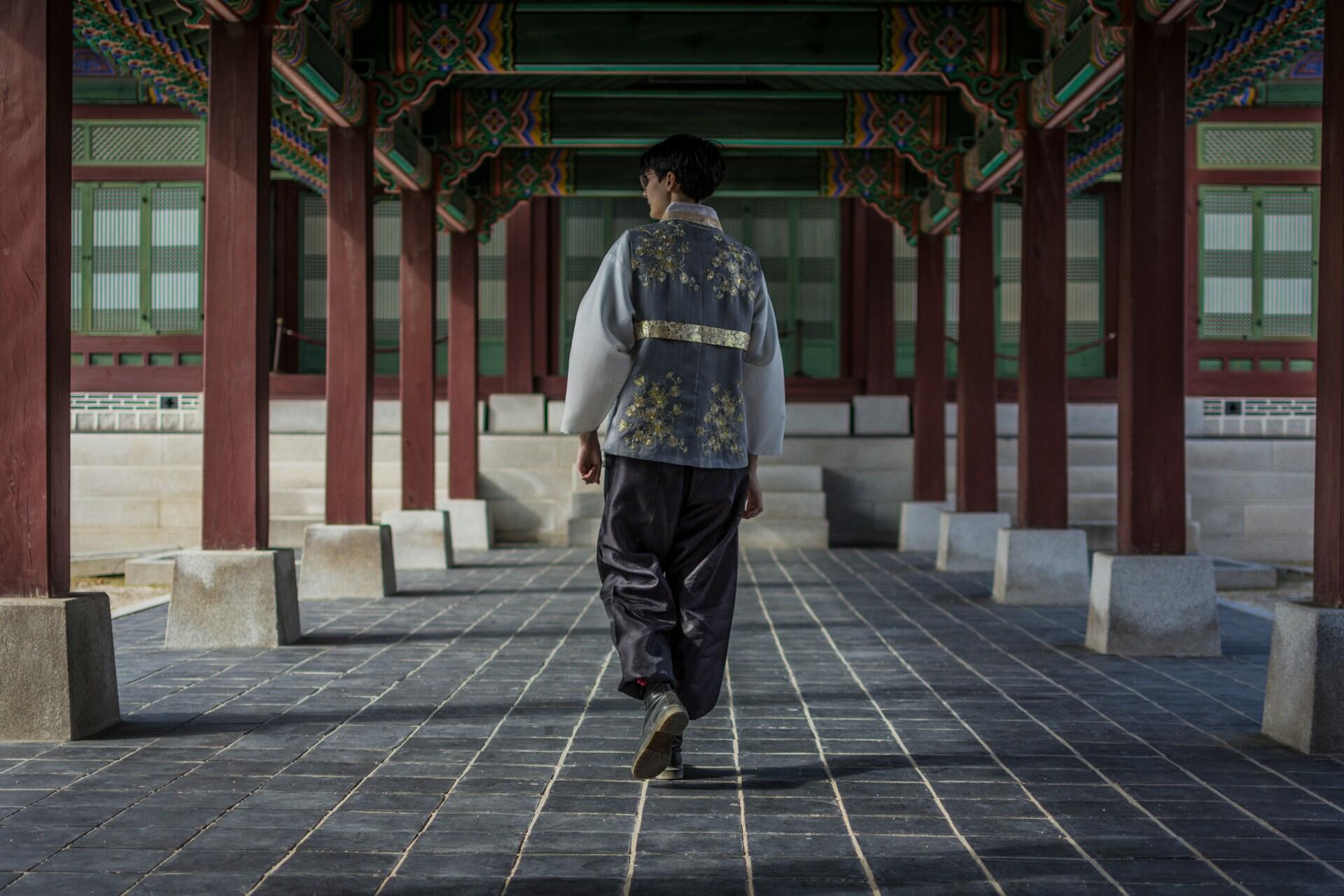 A young man walks along a sheltered walkway wearing a traditional Korean hanbok. The mood is solemn and reflective.