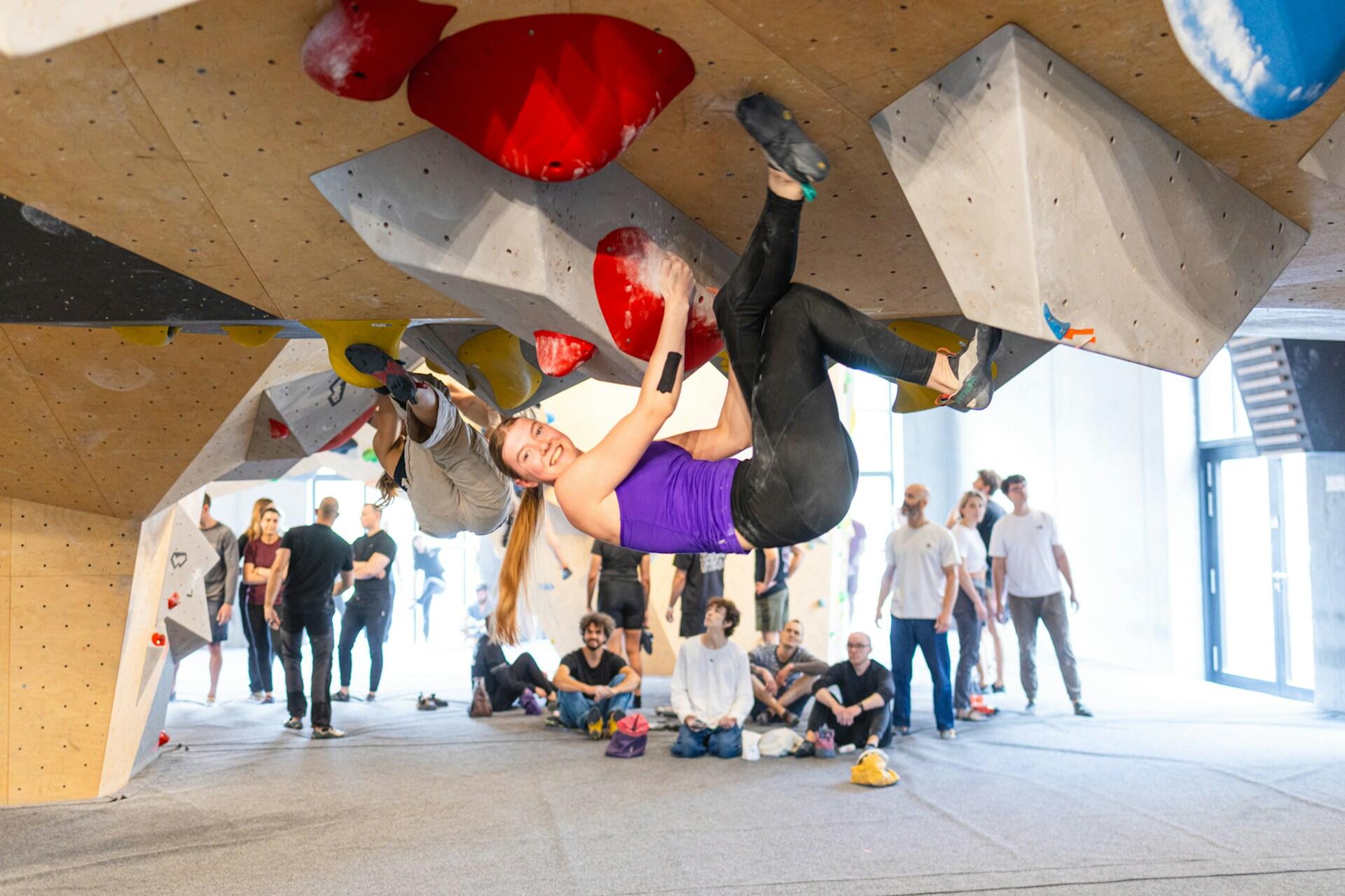 Woman bouldering on an overhanging indoor wall while other climbers watch